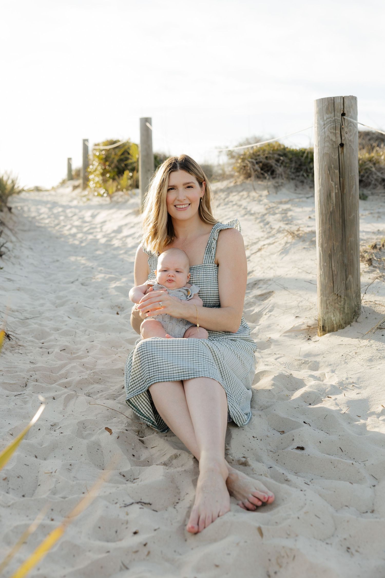Parent and baby sitting on sandy beach with wooden posts in background during golden hour.