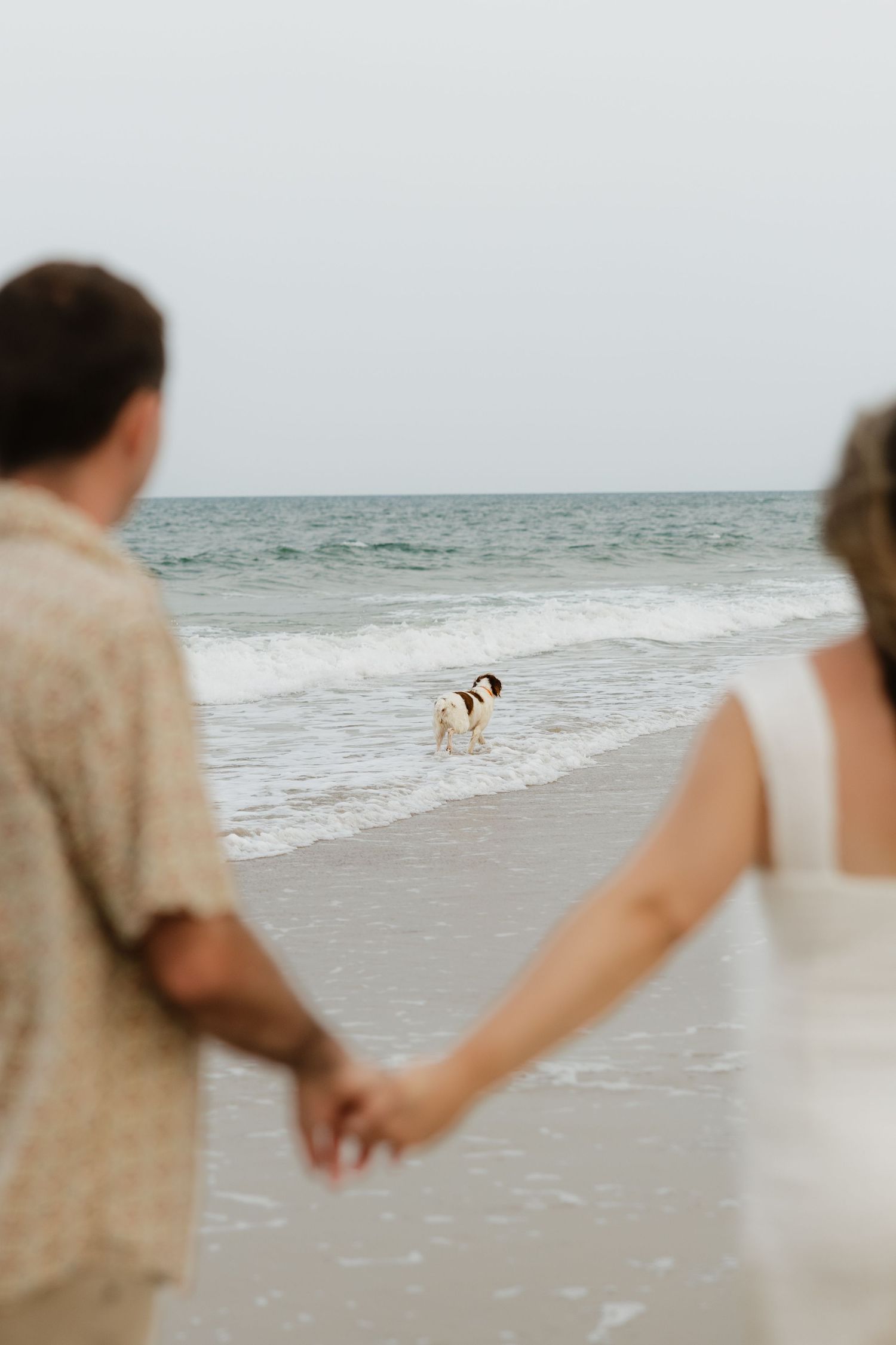 Couple walking hand in hand on beach with blurred ocean waves in background.