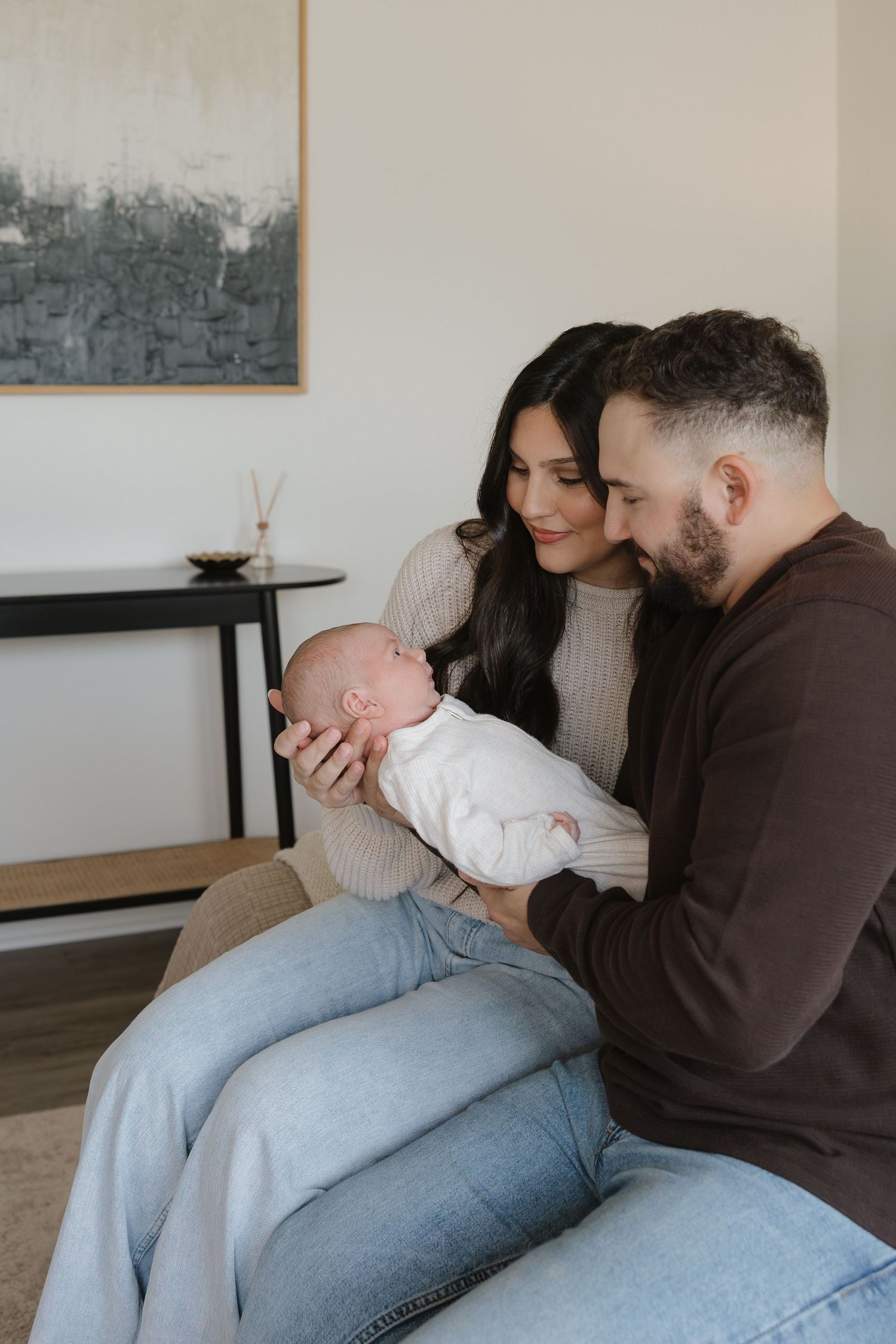 A couple in cozy sweaters sits together on a couch holding their newborn baby in a white onesie.