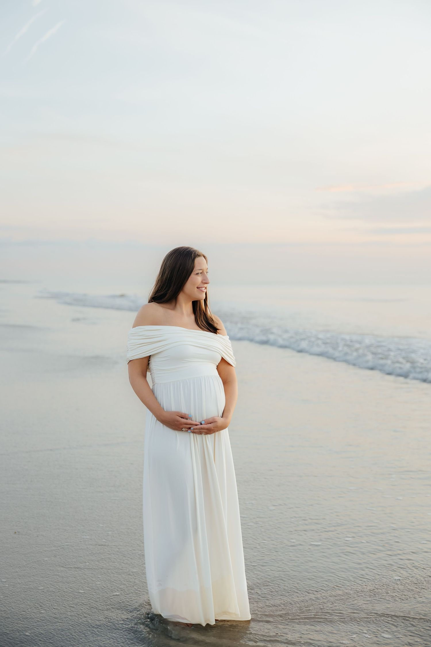 Pregnant woman in white off-shoulder gown stands at ocean's edge during sunset, cradling her baby bump.