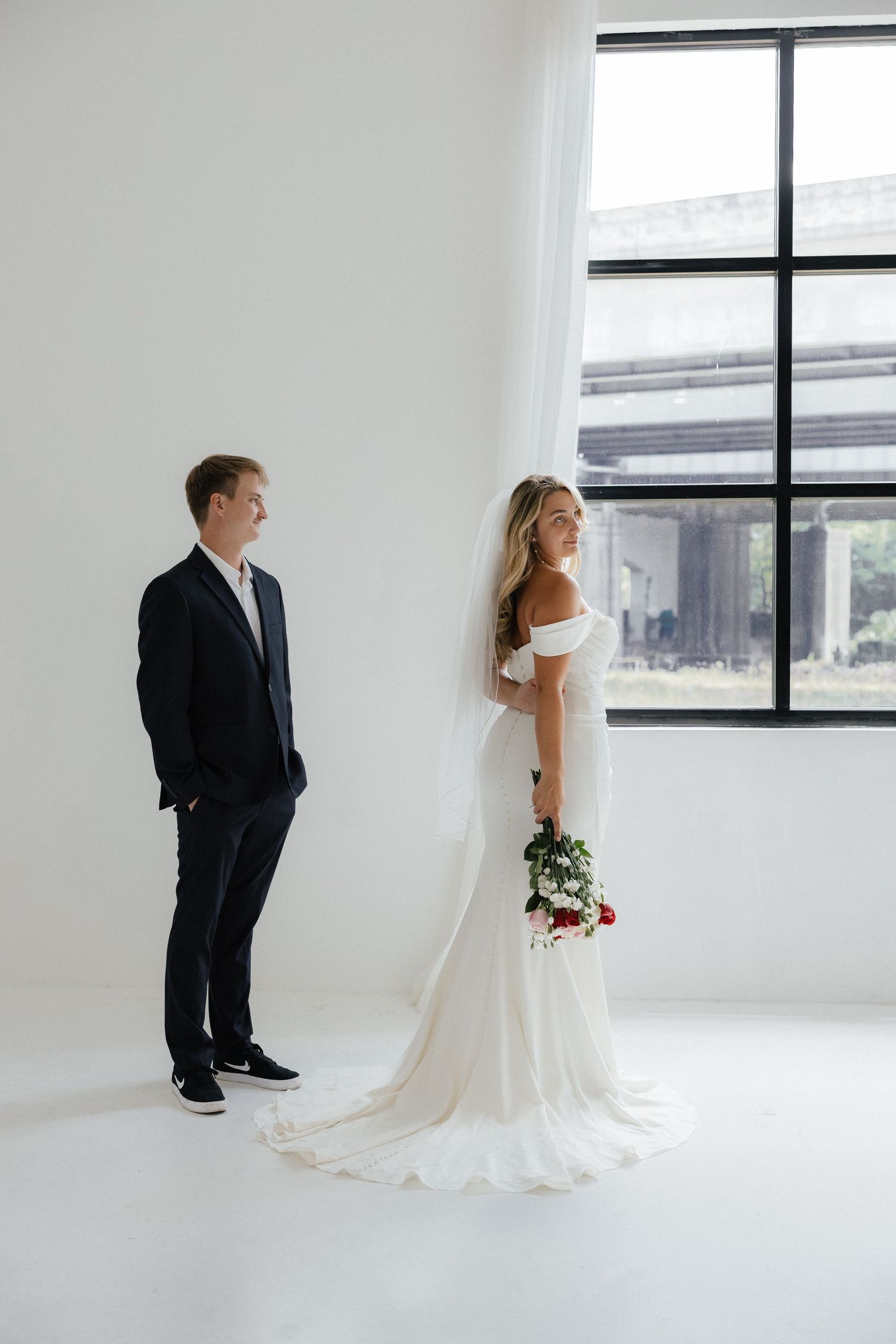 A bride and groom pose separately in a white room with large windows.