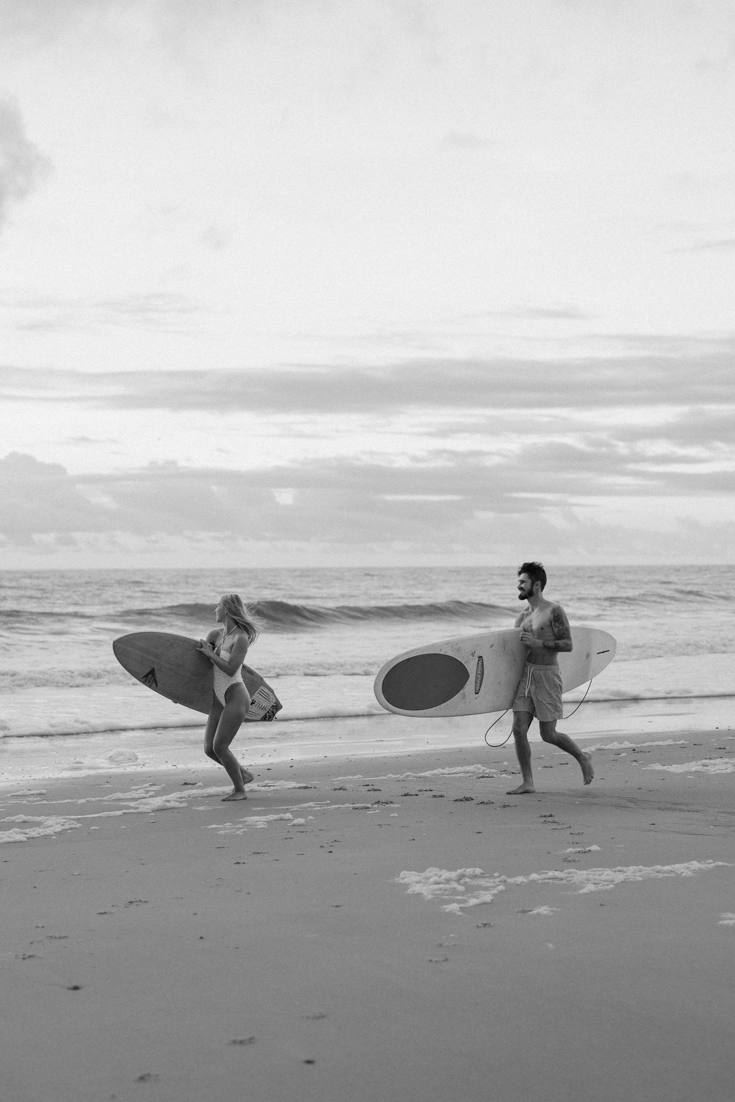 Two surfers carrying their boards along a misty beach shore in black and white.