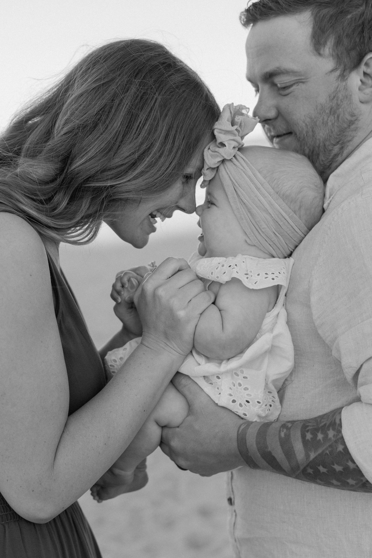 Black and white photo of smiling parents on a beach gently touching foreheads with their baby girl in a lace outfit and bow headband.
