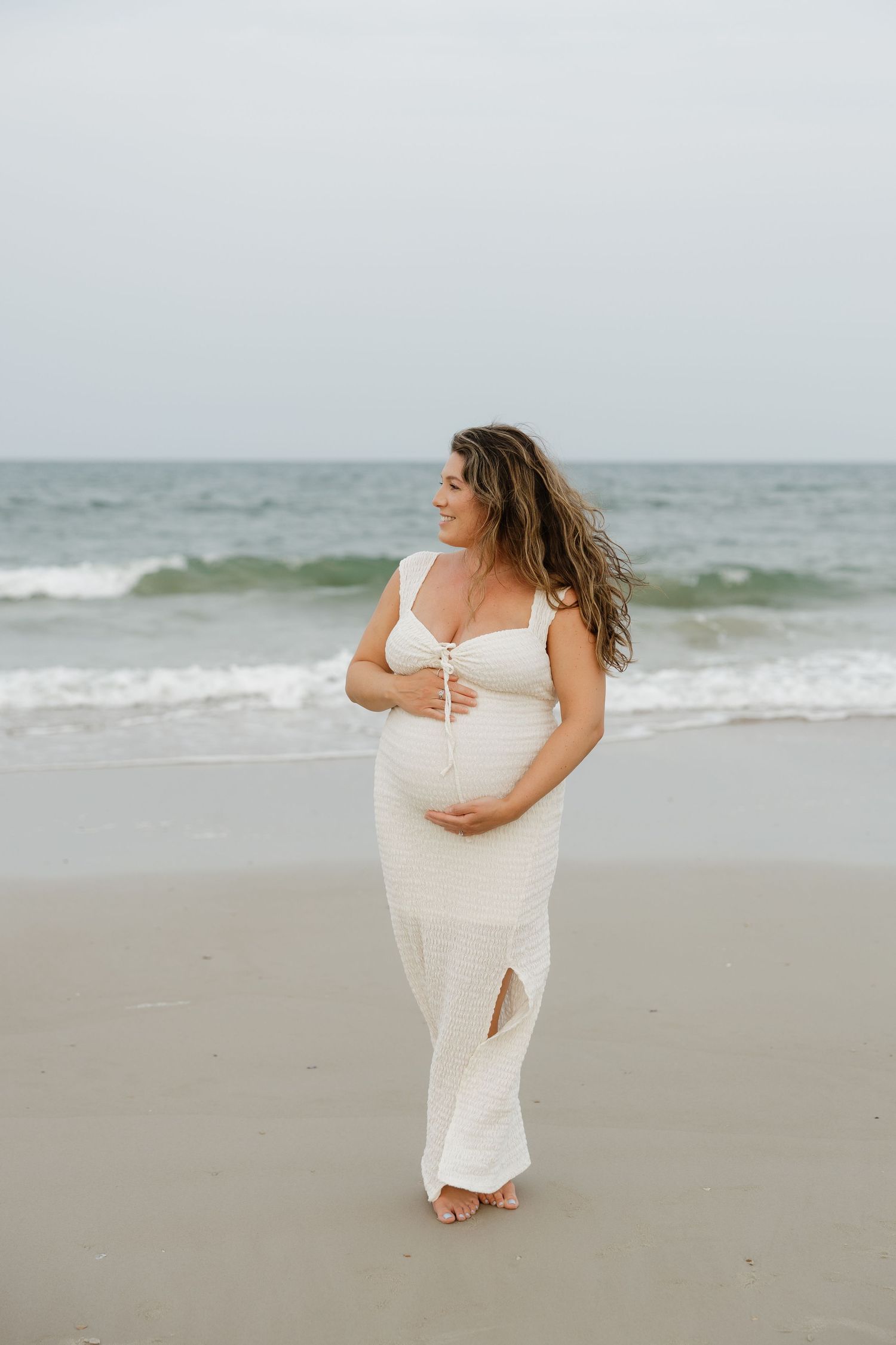 A pregnant woman in a white dress poses serenely on a beach during a maternity photoshoot.
