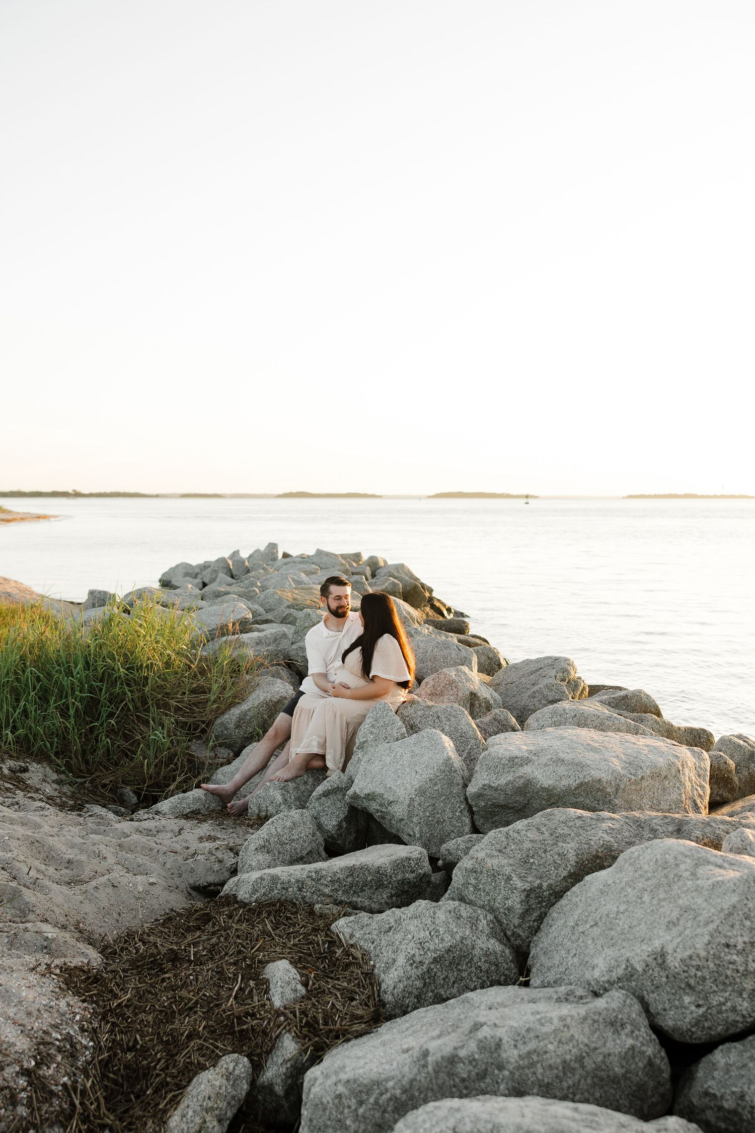 Couple sitting close together on large coastal rocks at sunset, with calm water and a grassy shoreline stretching toward the horizon.
