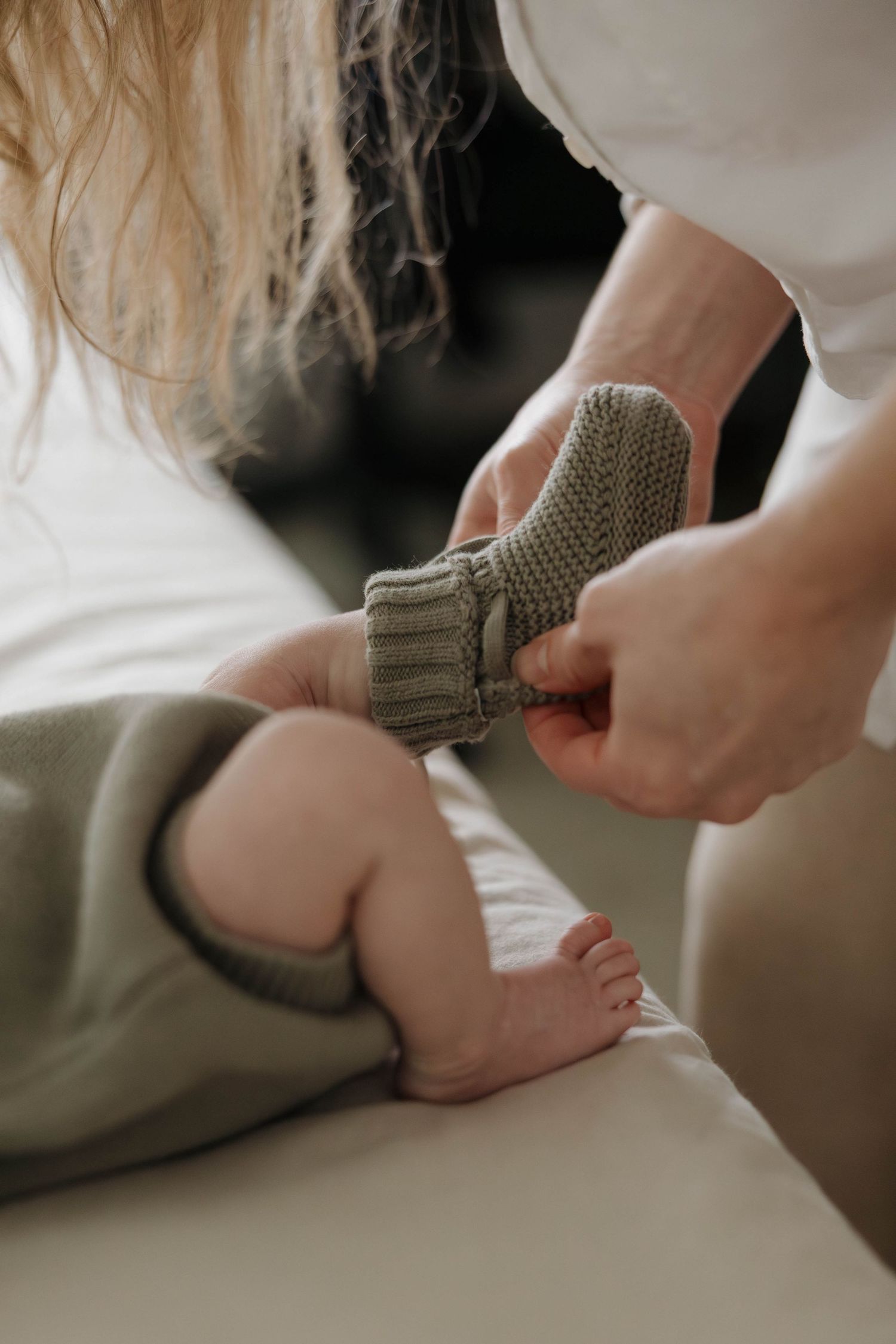 Close-up view of putting on a small gray knit sock or bootie on tiny feet against white bedding.