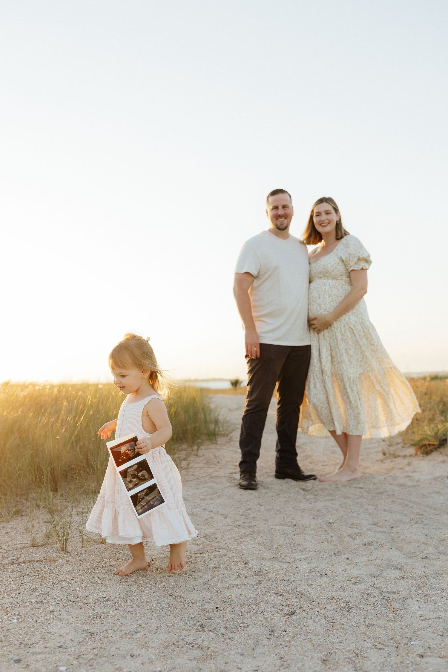 Family poses on sandy beach in Fort Clinch State Park during sunset while small child holds ultrasound photo.