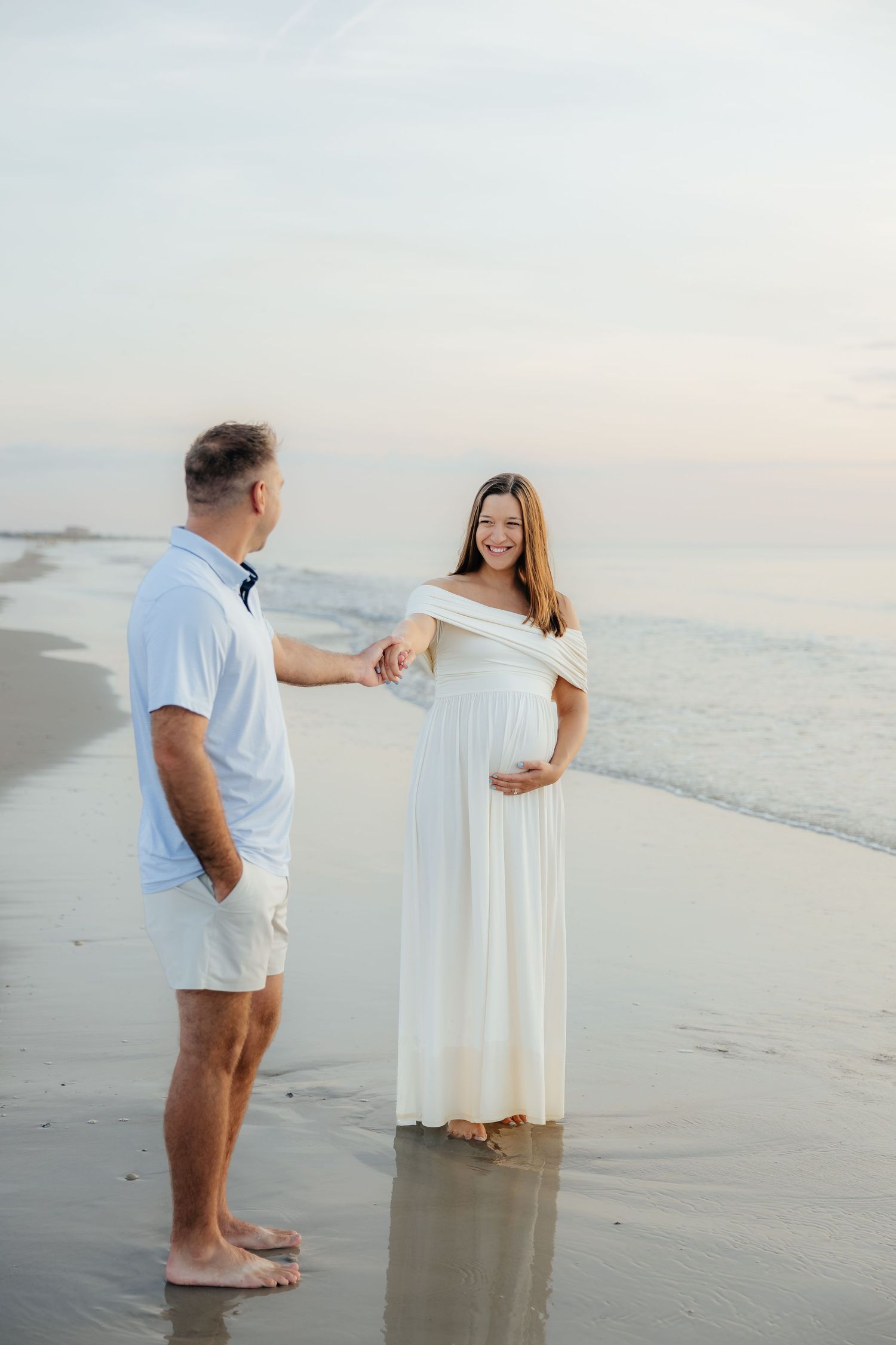 Pregnant woman in white dress holds hands with man on beach at sunset, smiling joyfully during maternity photoshoot.