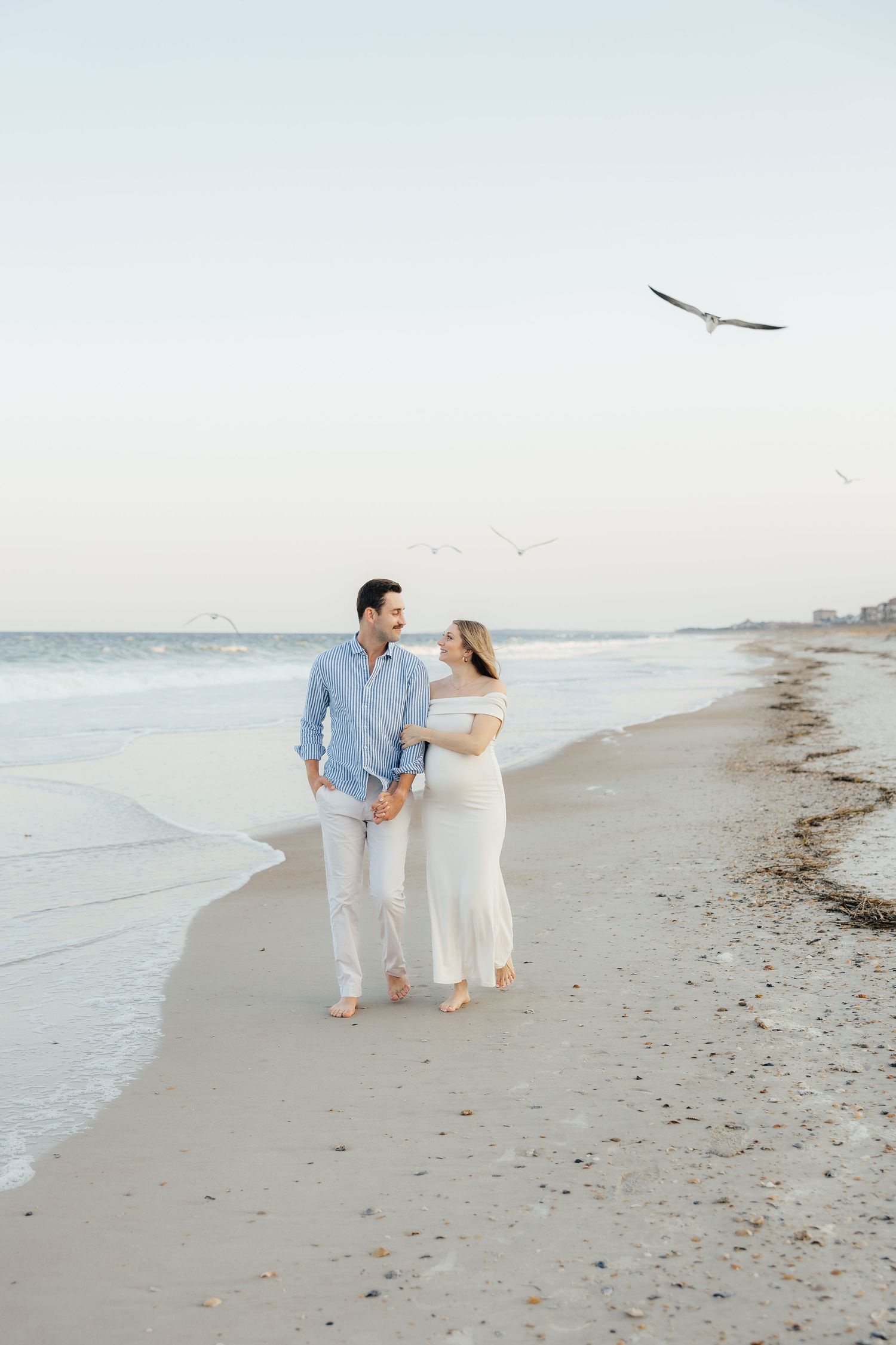 A couple walks hand in hand along a serene beach shoreline during sunset in light colored outfits.