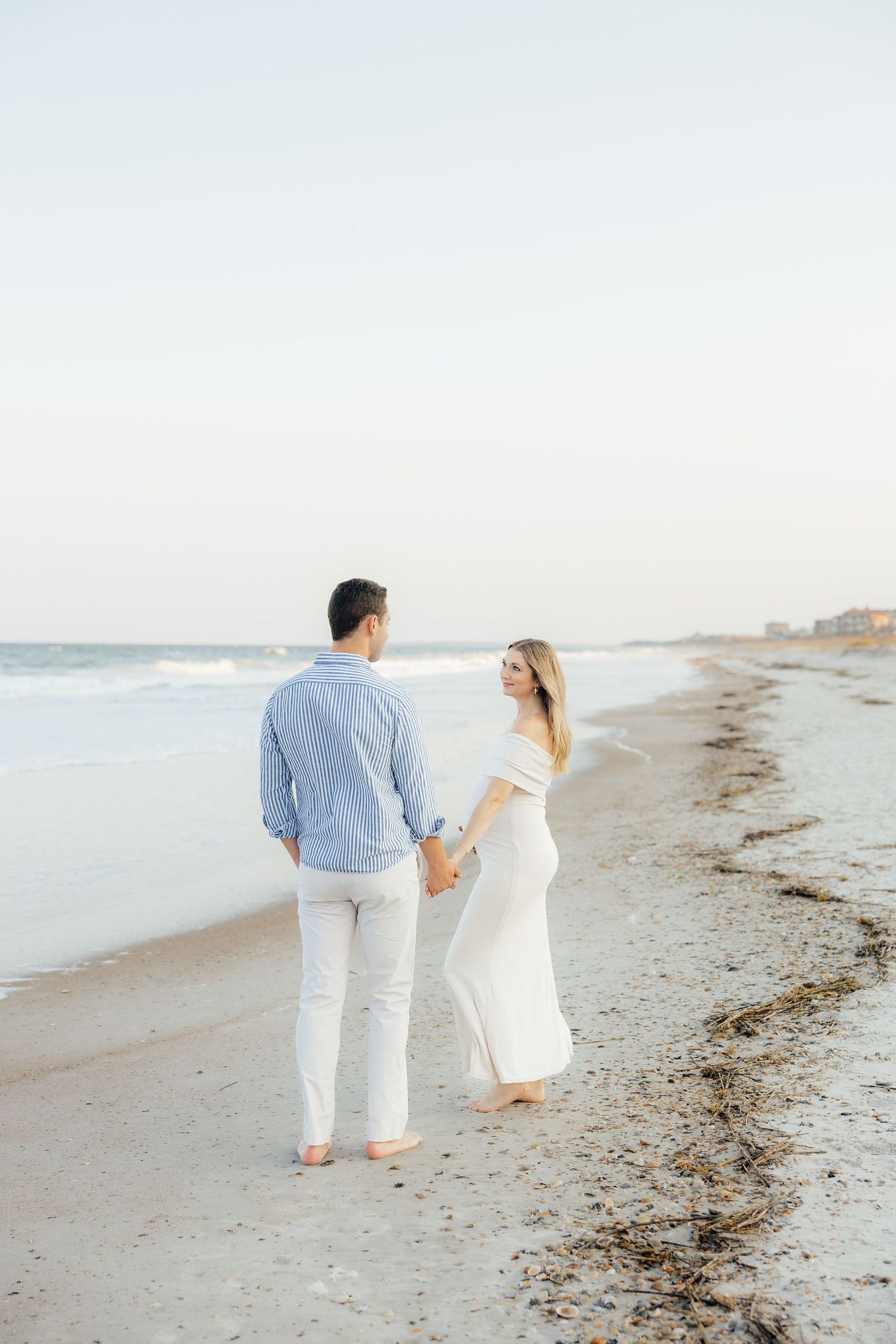 A couple walks barefoot along a serene beach at sunset wearing coordinated light blue and white outfits.