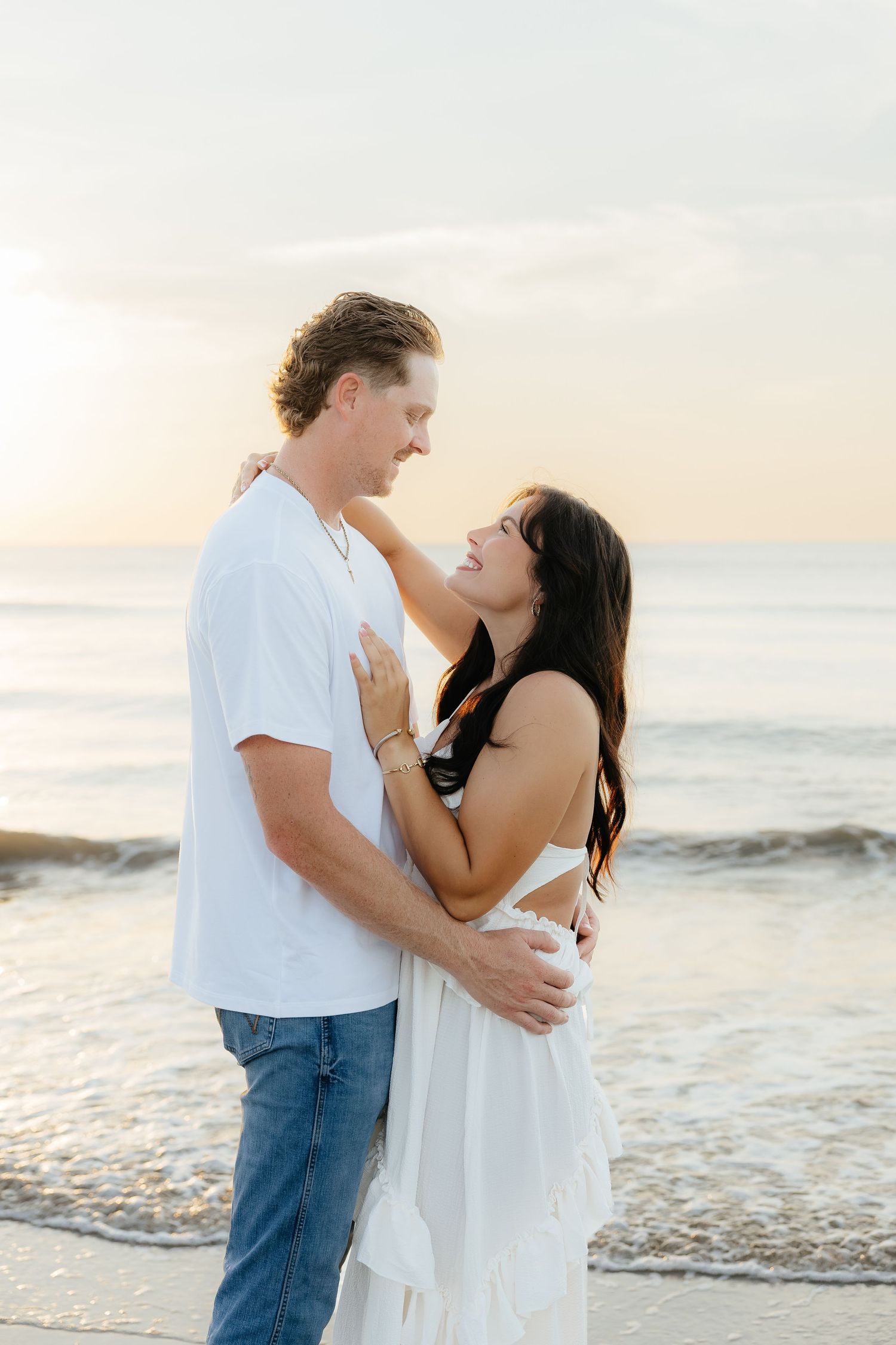 A couple embraces and gazes at each other romantically on a beach at sunset, woman in white dress, man in white tee.