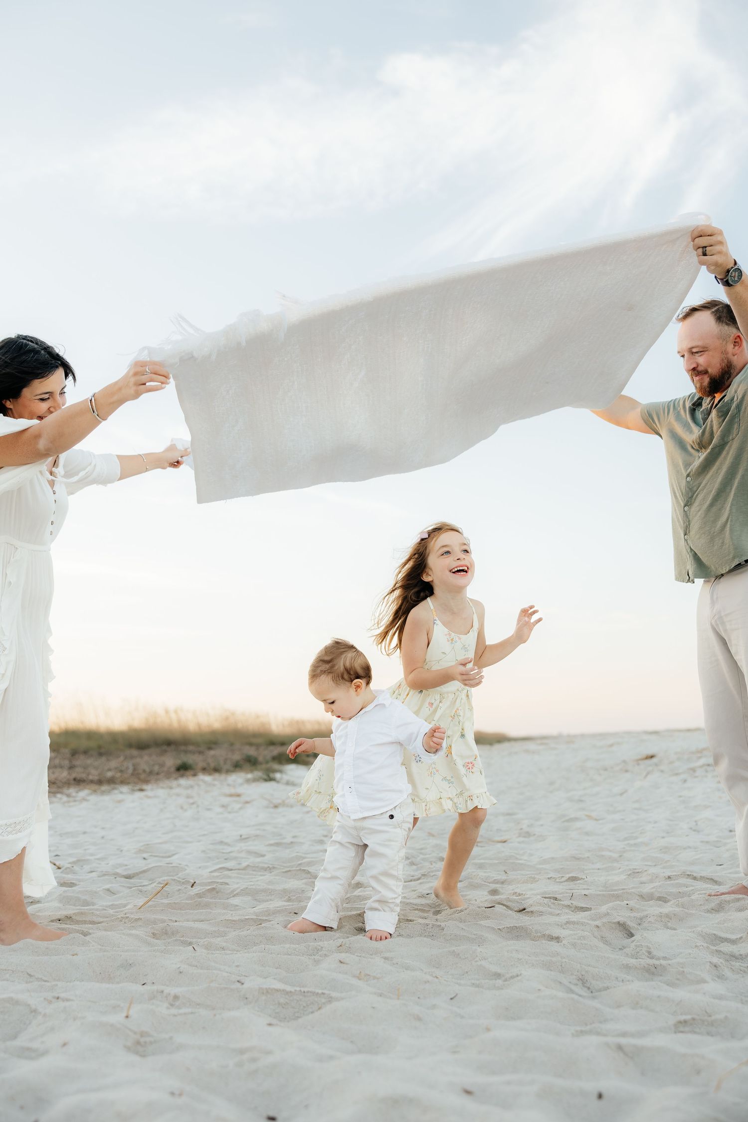 Family in white clothing playing together on a sandy beach at sunset, creating a joyful and candid moment.