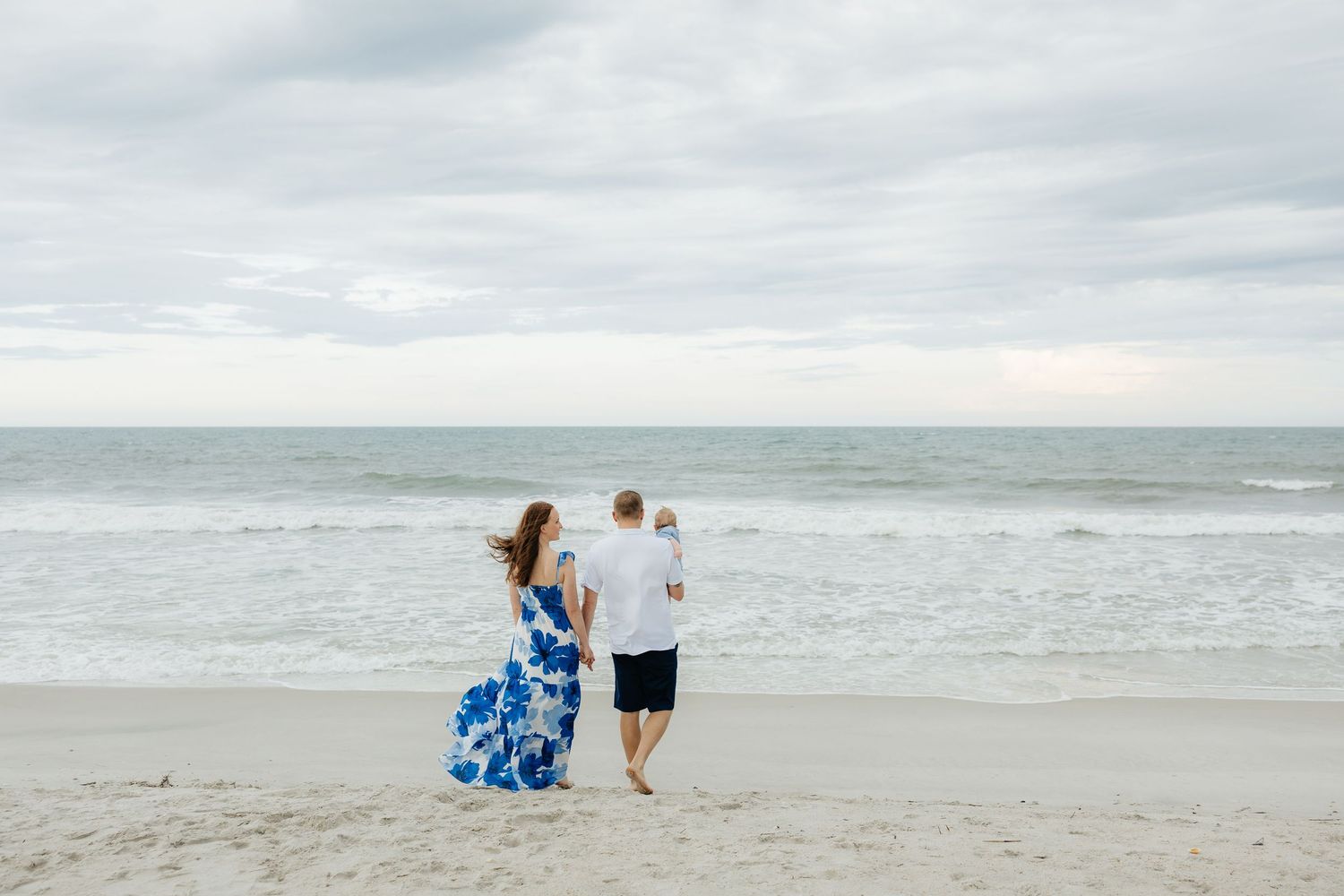 Couple walking along ocean shoreline with waves and cloudy sky in background.