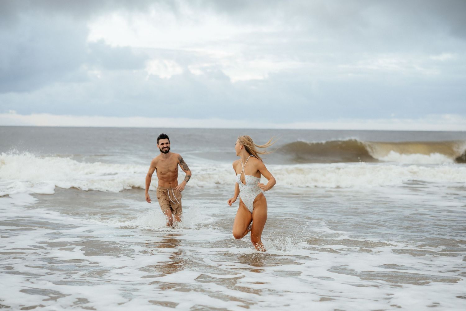 Couple playing in ocean waves on cloudy beach day.