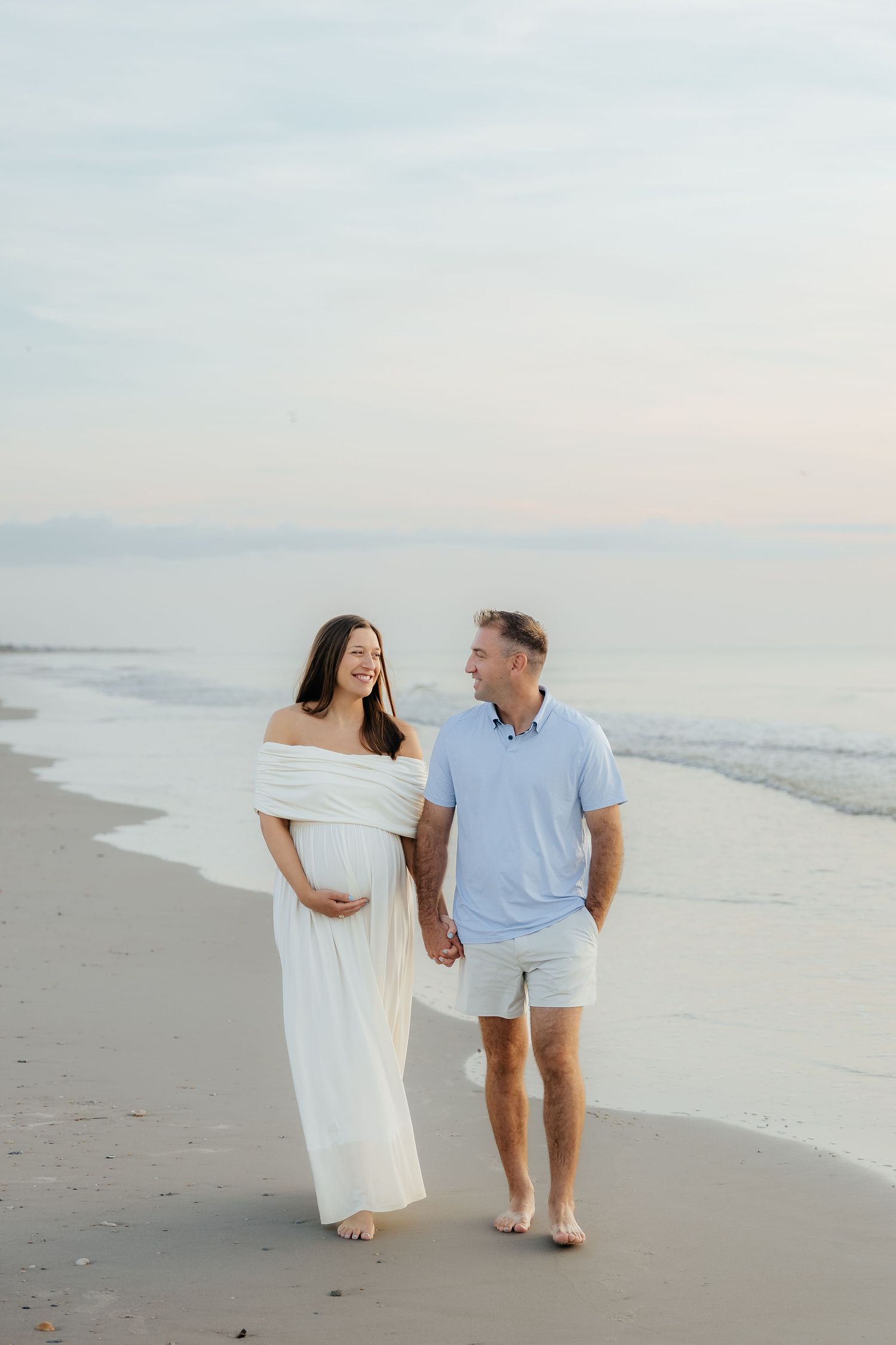 Pregnant couple walks hand-in-hand on a serene beach at sunset, woman in white off-shoulder gown cradling her baby bump.
