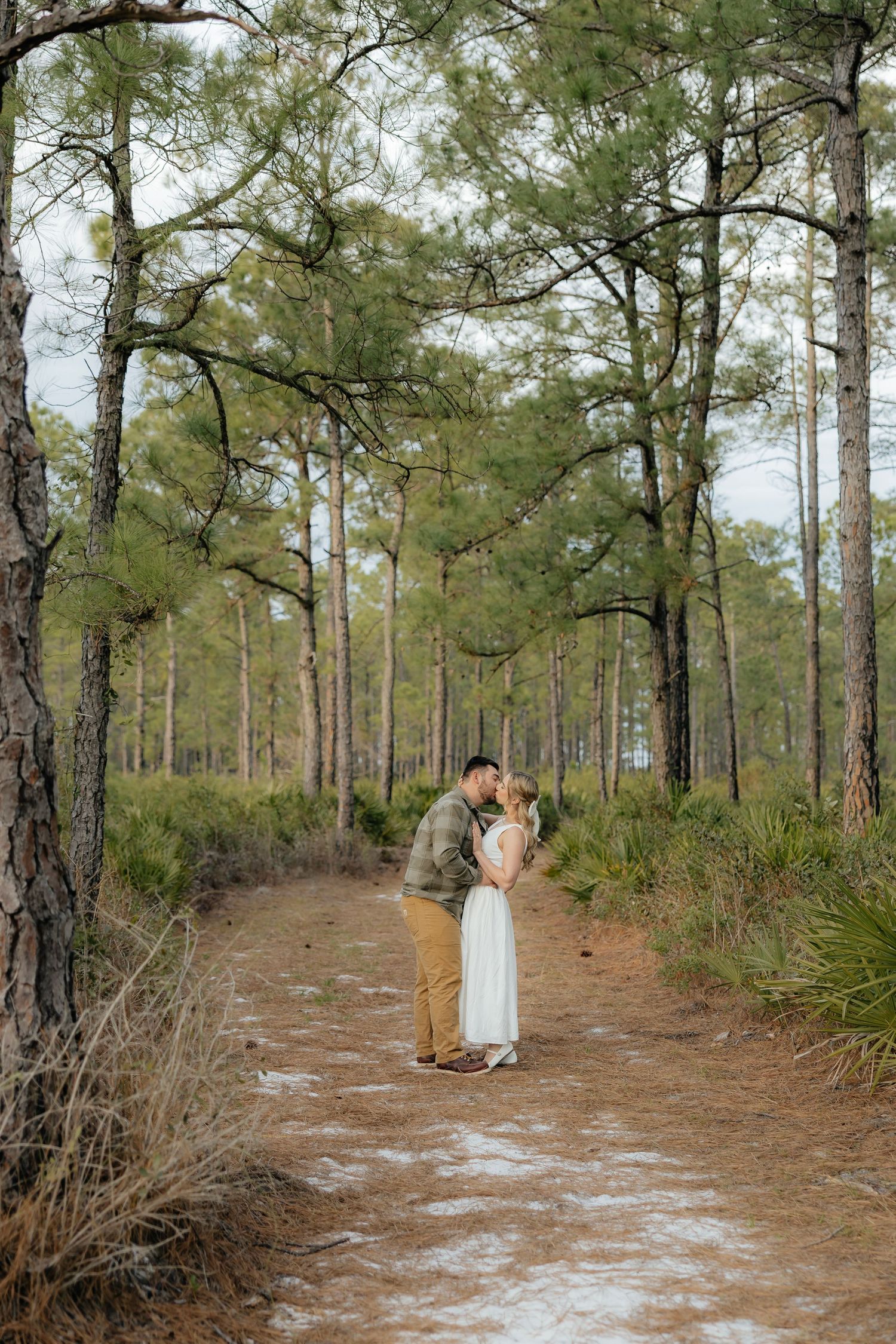 Couple in an olive shirt, tan pants and white dress share a kiss on a sandy forest trail lined with tall pine trees and palmetto.