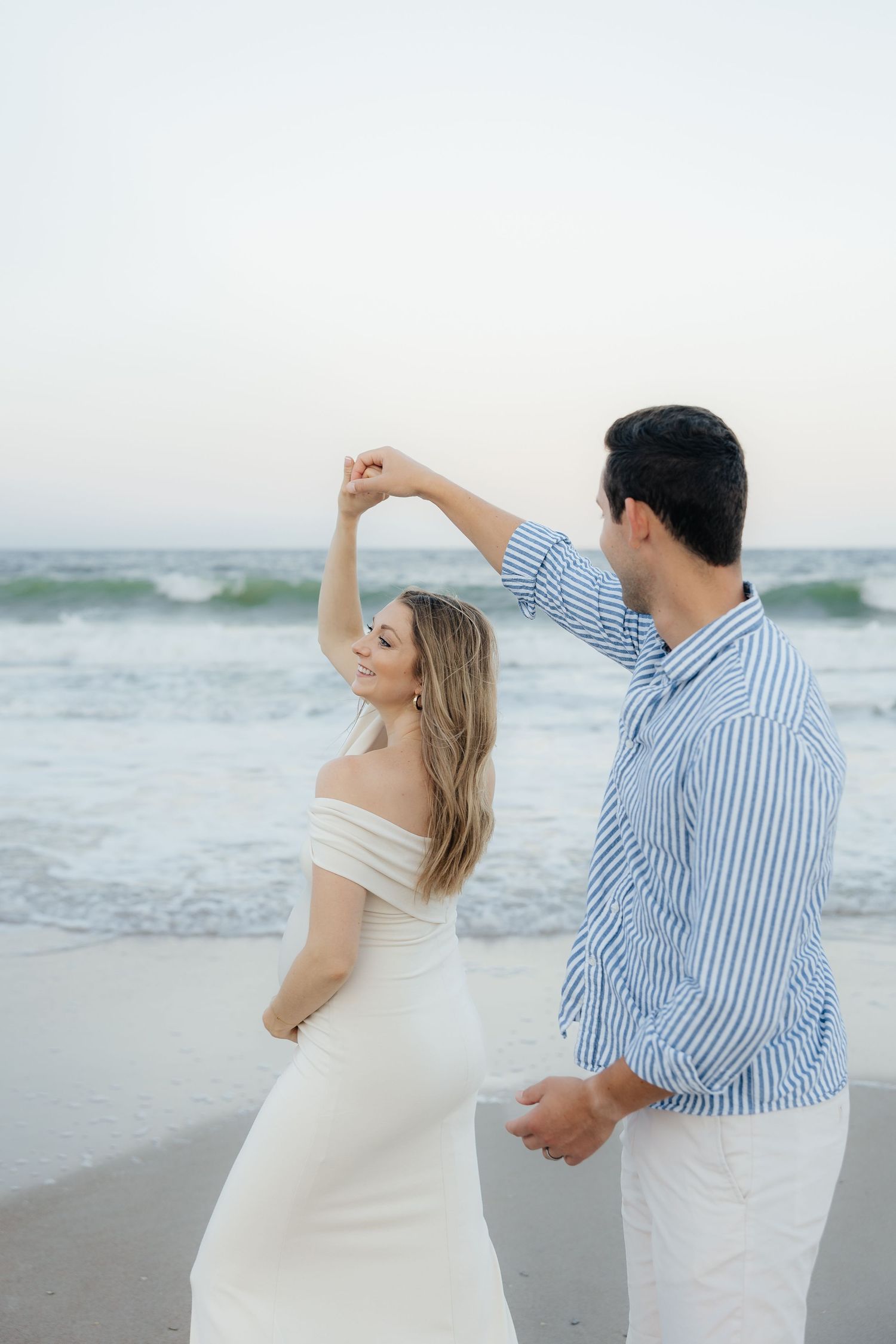 A couple dances together on a beach at sunset with ocean waves in the background.