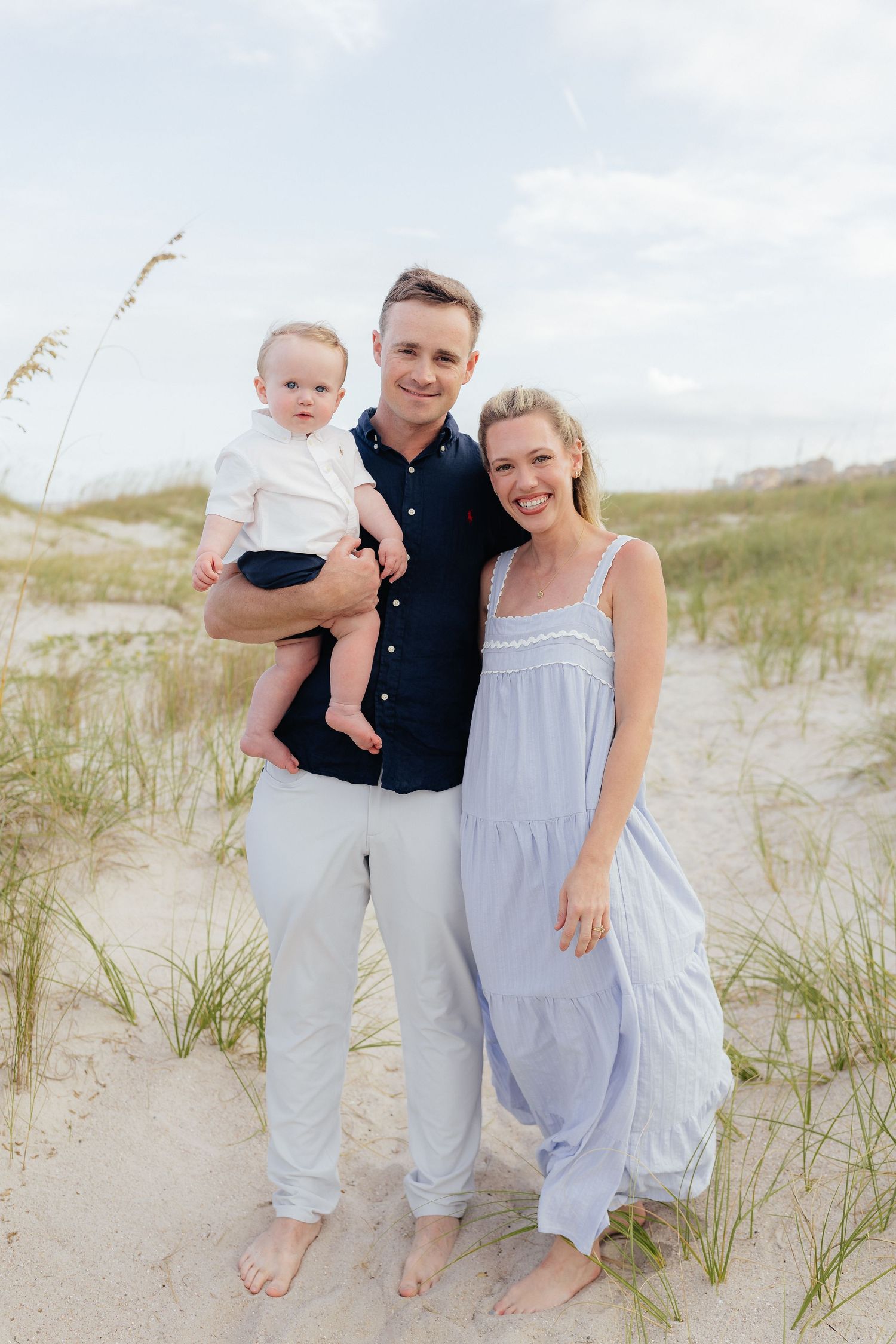 Family of three dressed in coastal casual attire pose together on sandy beach dunes.