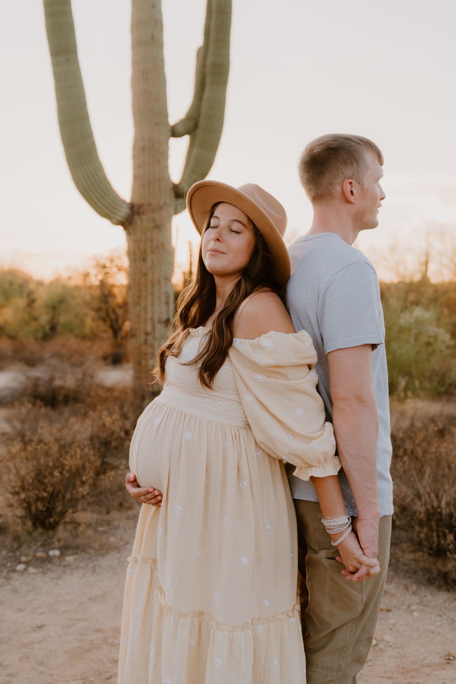 Expectant couple poses against desert backdrop with tall saguaro cactus during golden hour.