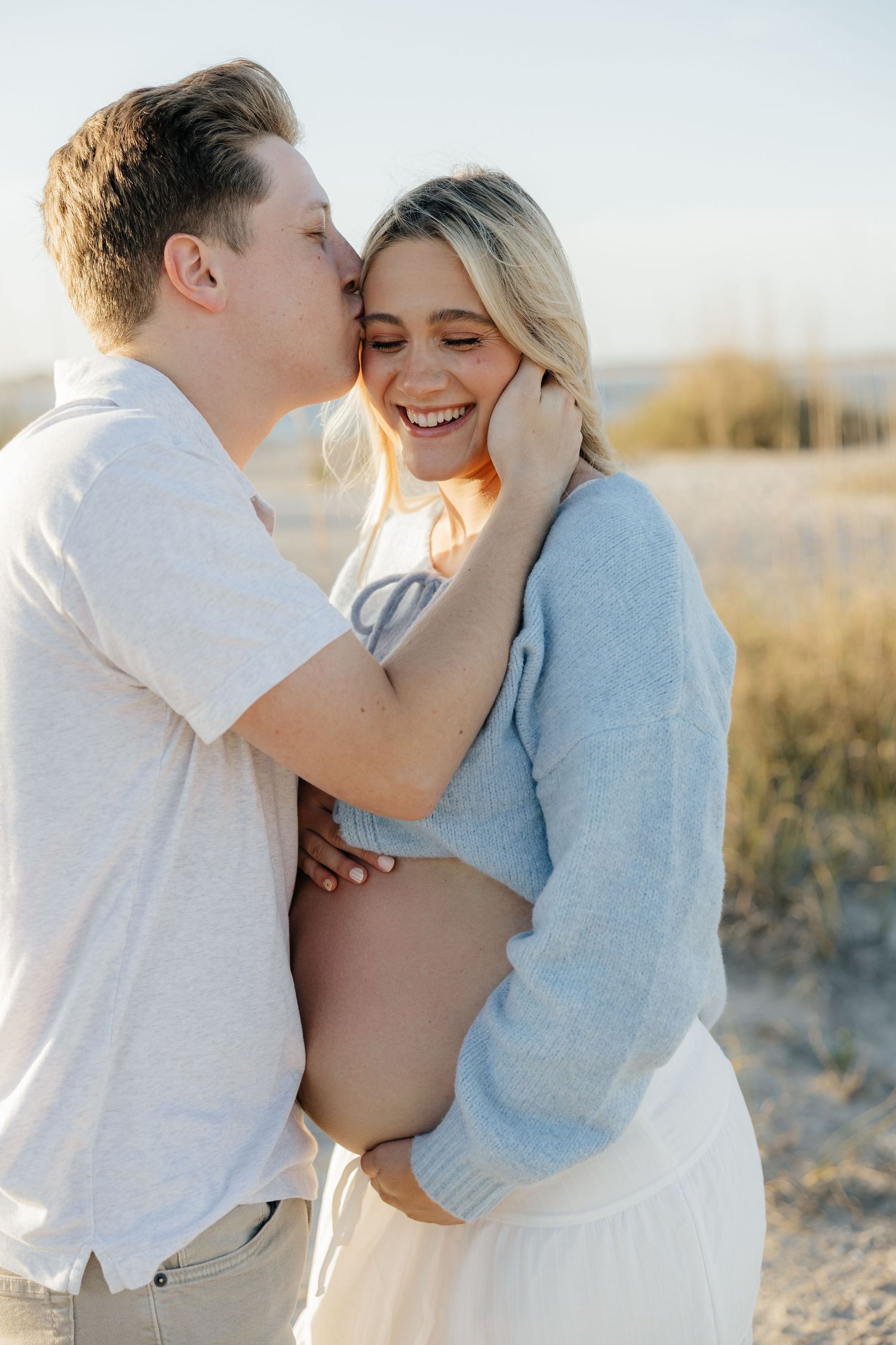 A couple shares tender moments on a beach at sunset in soft pastel tones, wearing white and blue casual attire.