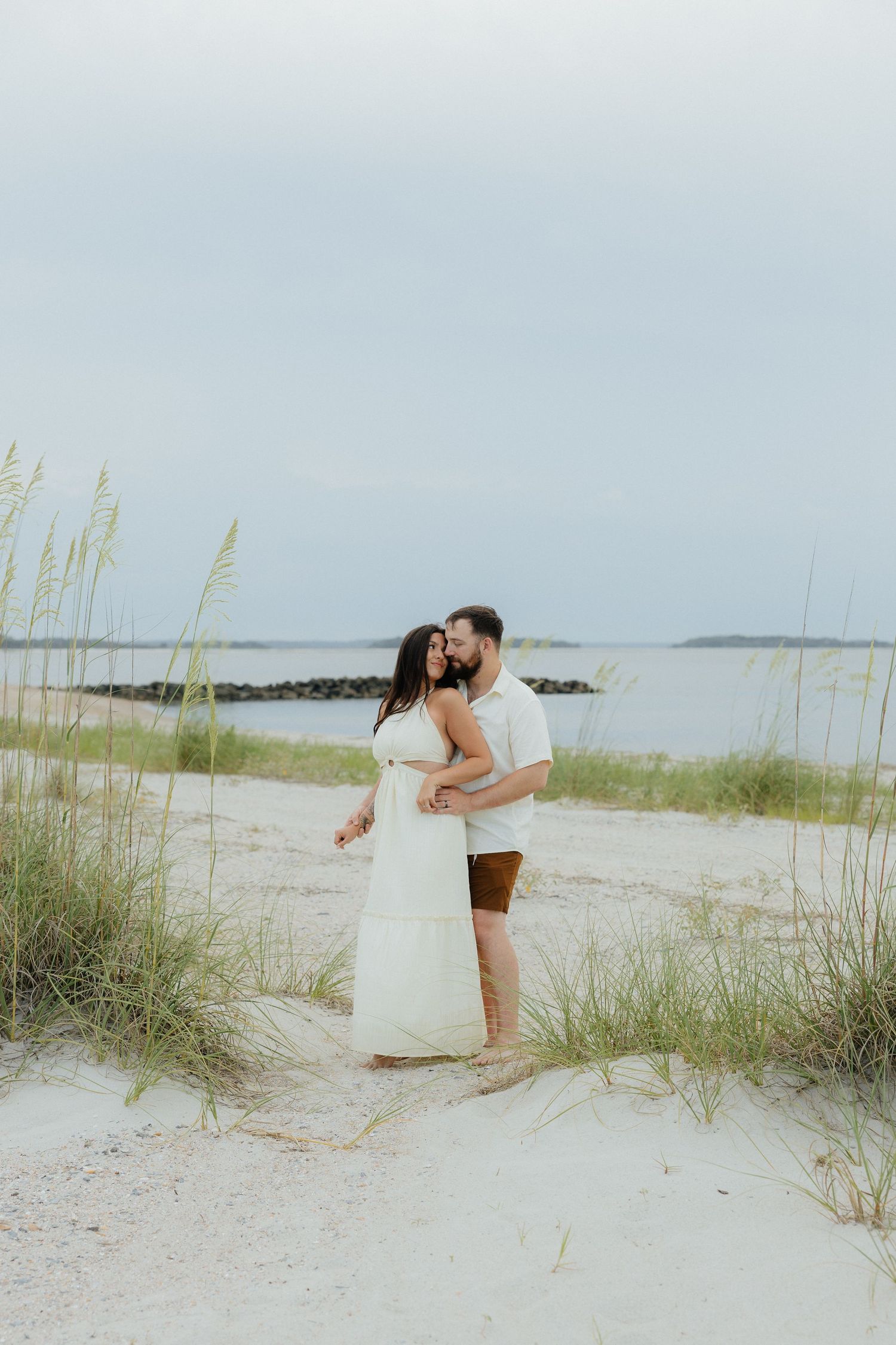 A couple embracing among beach grass dunes with the ocean in the background.