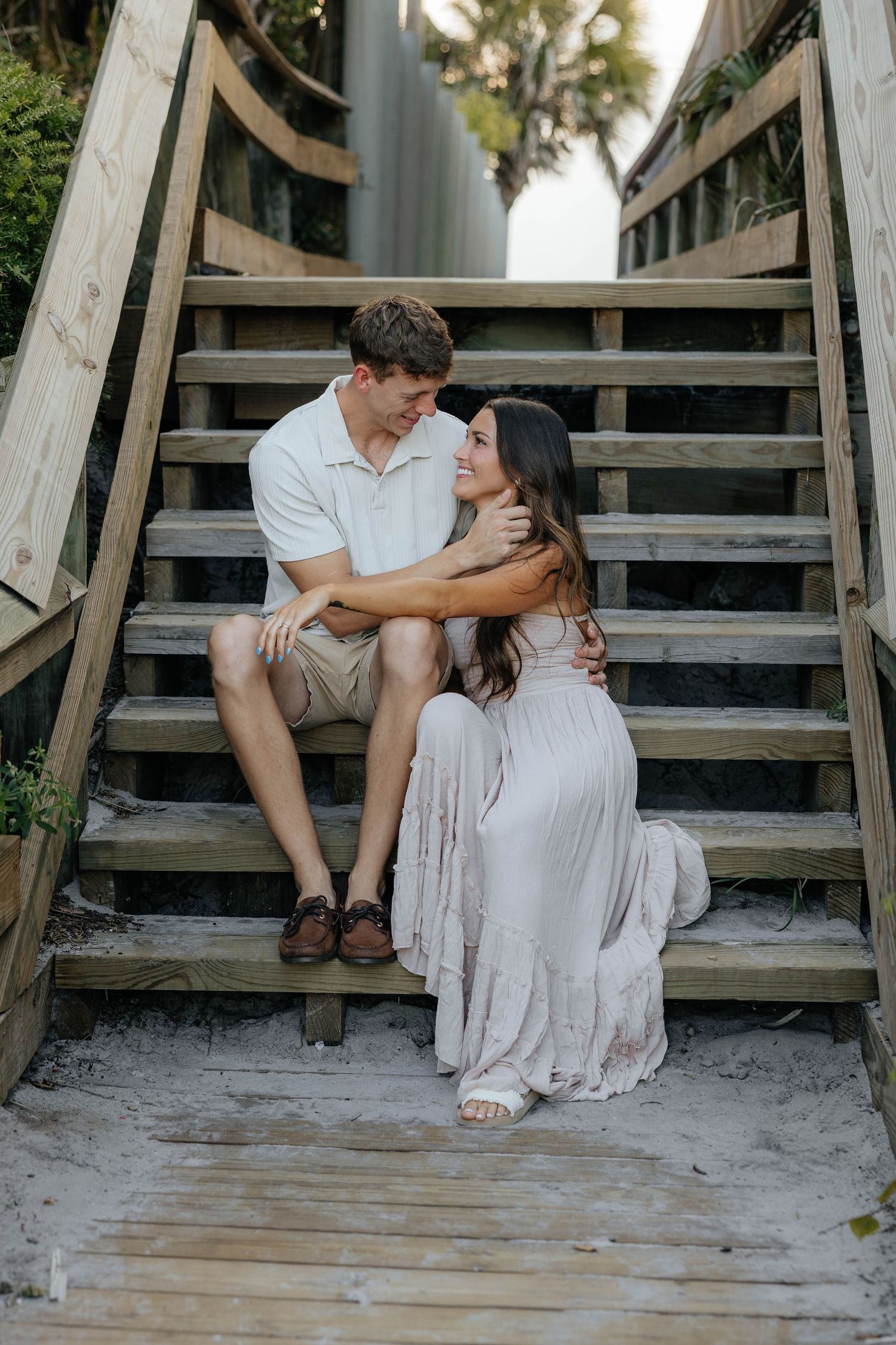A romantic couple sits together on wooden beach boardwalk steps in casual summer attire.