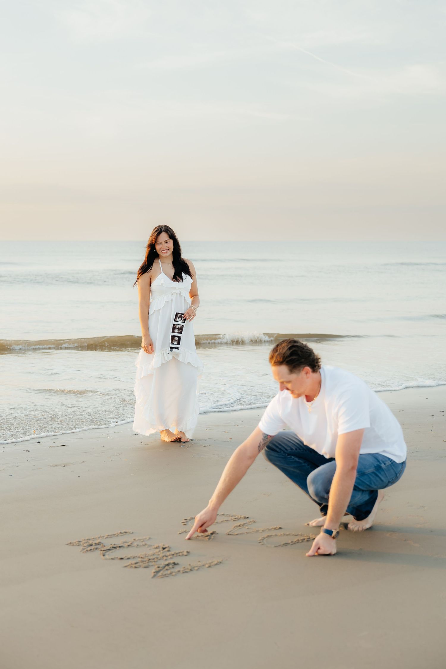 Couple at beach, man writing in sand, pregnant woman in white dress holding ultrasound photos, maternity announcement.