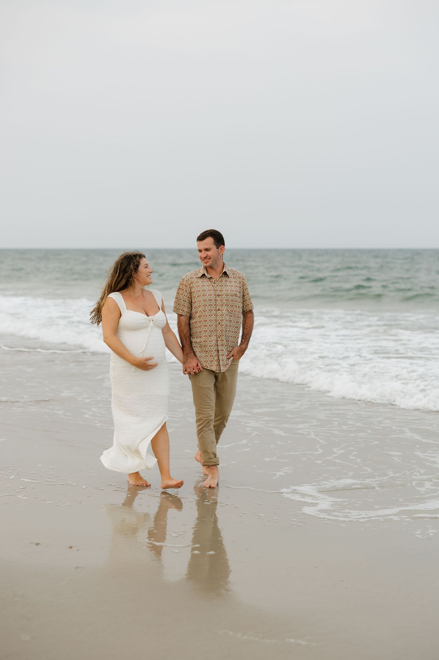 Happy couple playfully walks along the shoreline during a beach photo session.