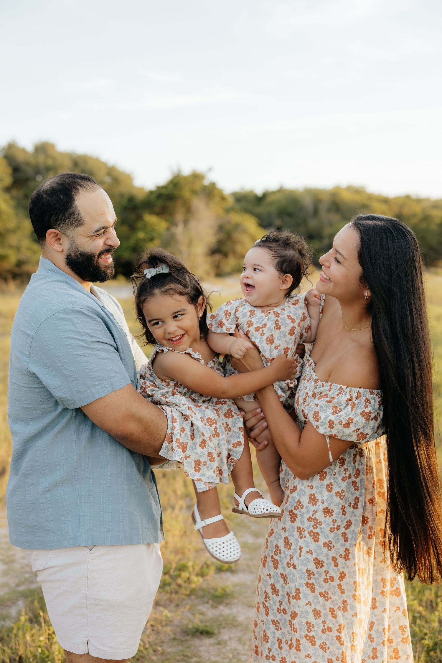 A happy family of four poses outdoors, with parents holding two young daughters in matching floral dresses in a sunny field.