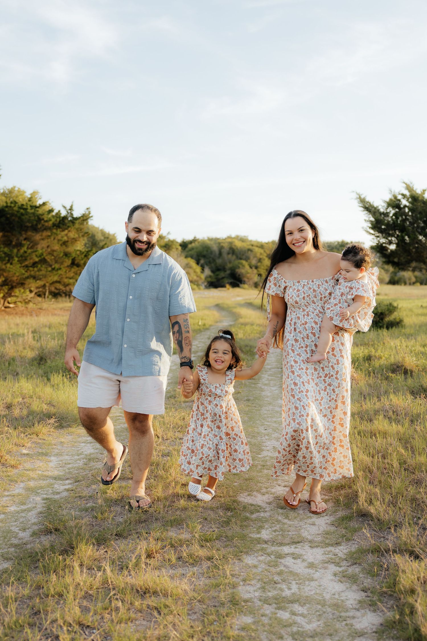 Smiling family of four in matching floral and blue outfits walking hand in hand along a grassy path during golden hour.