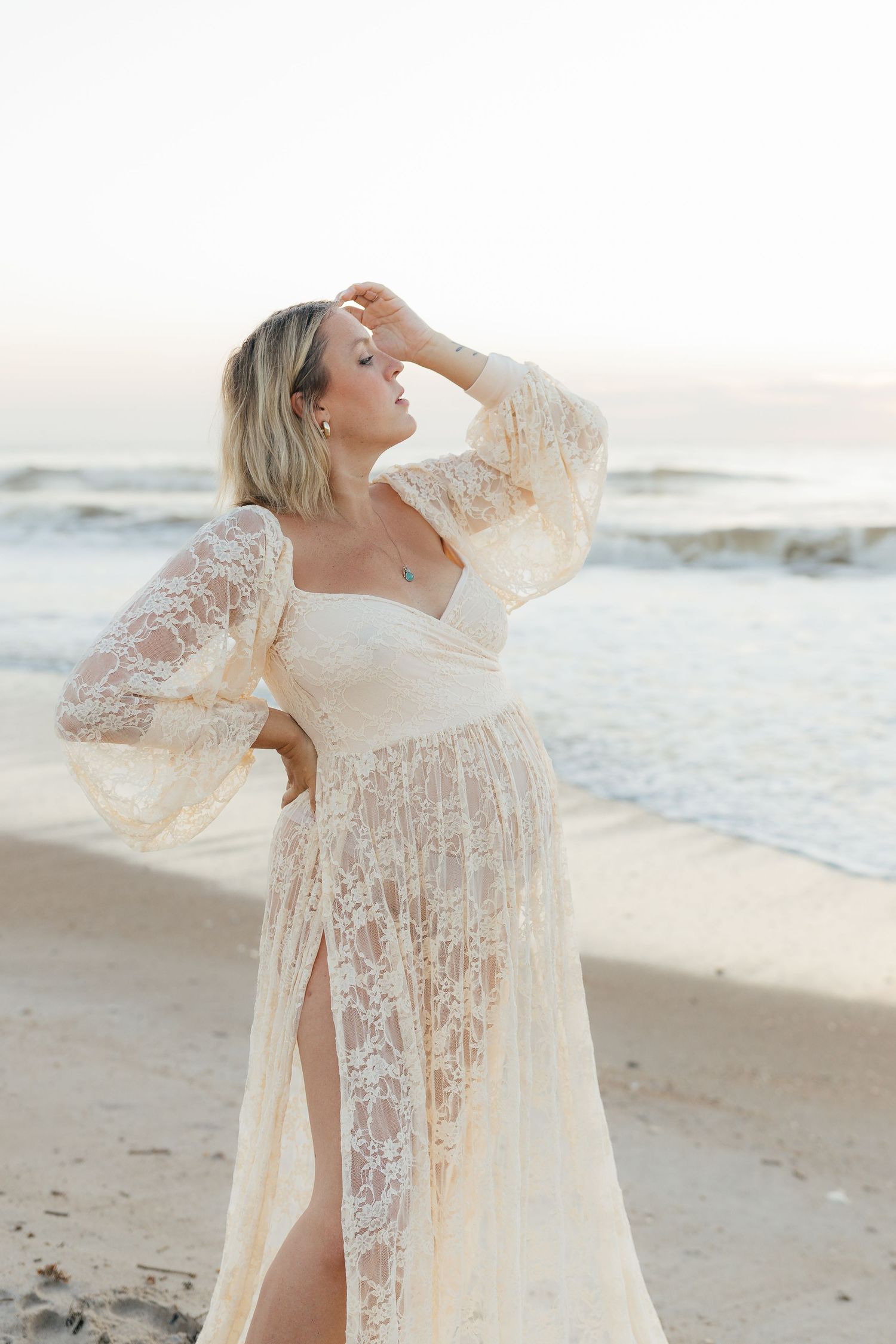 Woman in flowing lace dress poses elegantly on beach at golden hour.