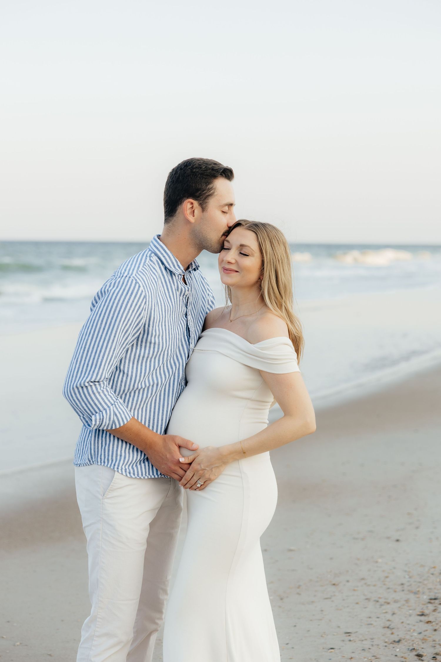 A romantic couple embraces on a serene beach at sunset wearing coordinated white and blue outfits.