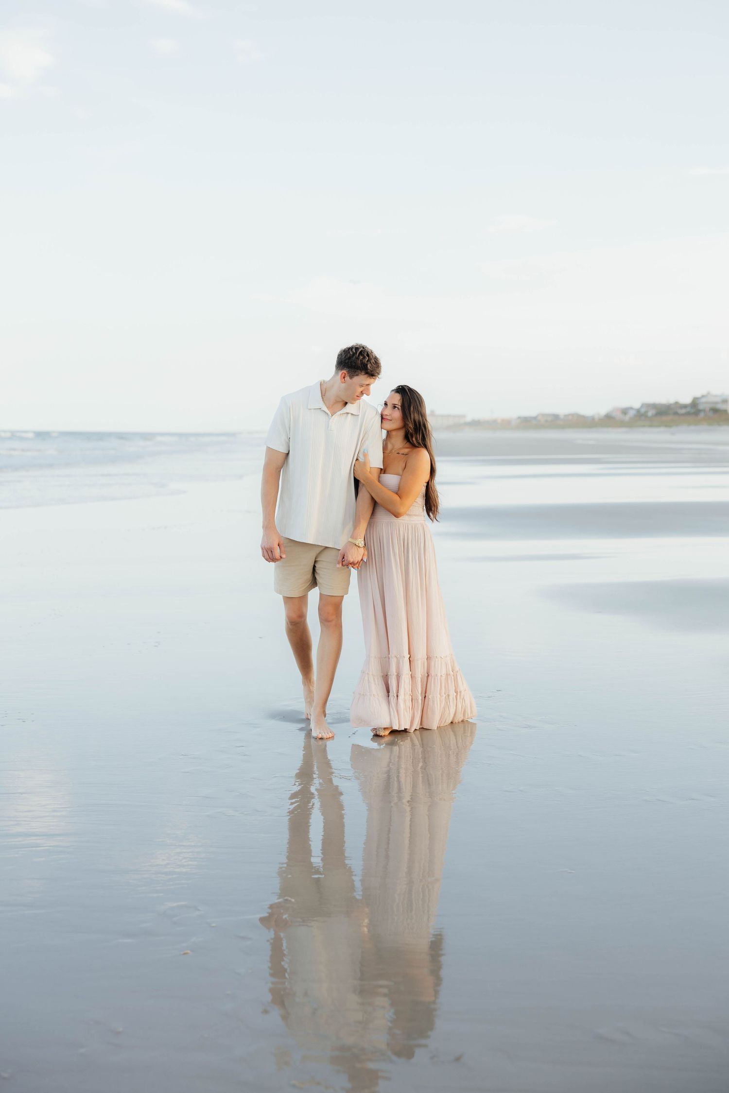Couple in light colored outfits walking barefoot along a tranquil beach shoreline at dusk with soft ocean reflections.