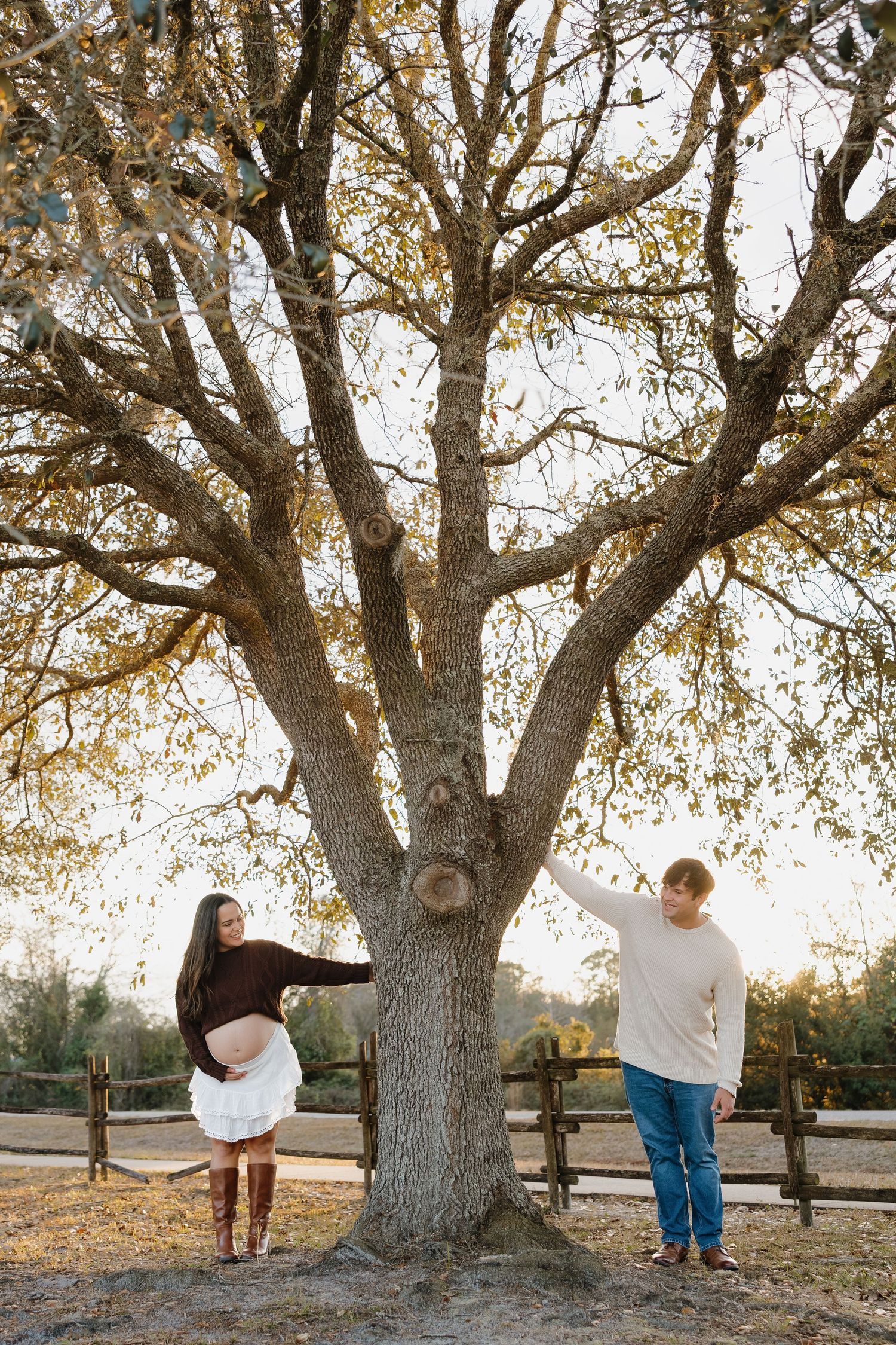 Pregnant woman in a white skirt and boots cradles her belly beside a man reaching toward a large tree in an autumn maternity photo.