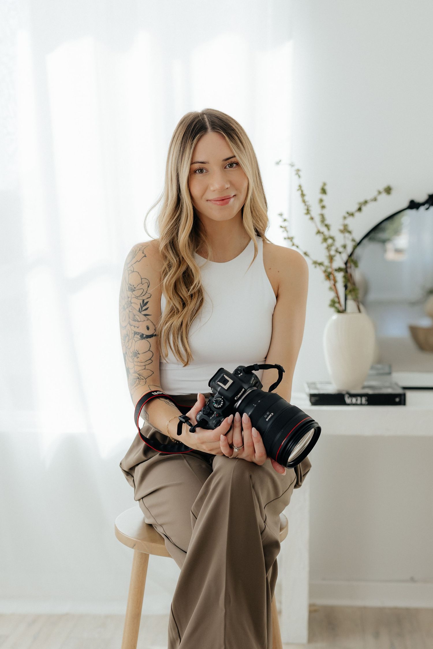 Photographer in white tank top with camera sitting on stool in minimalist studio.