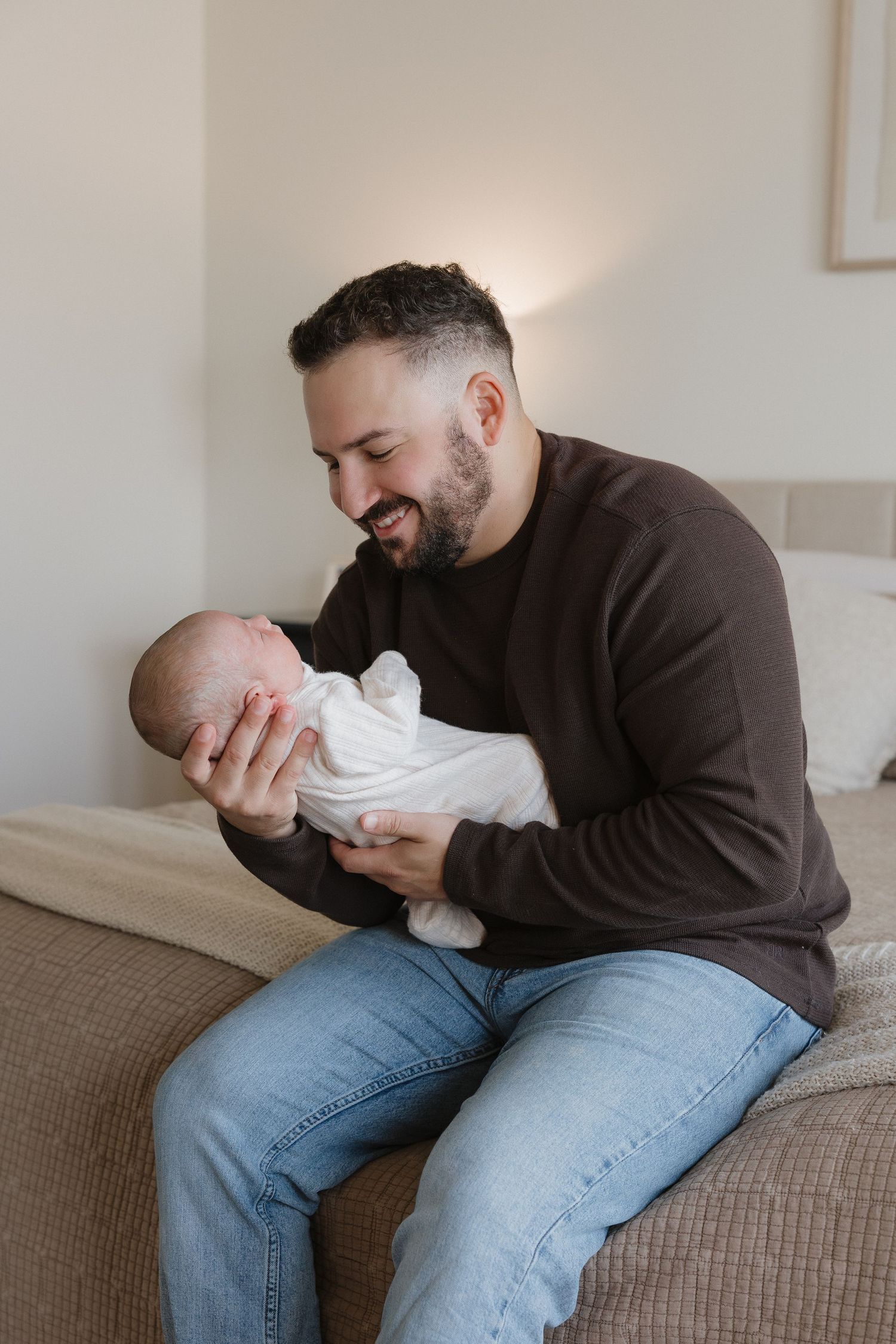 A parent in a brown sweater and jeans sits on a beige couch holding a baby in white clothing.