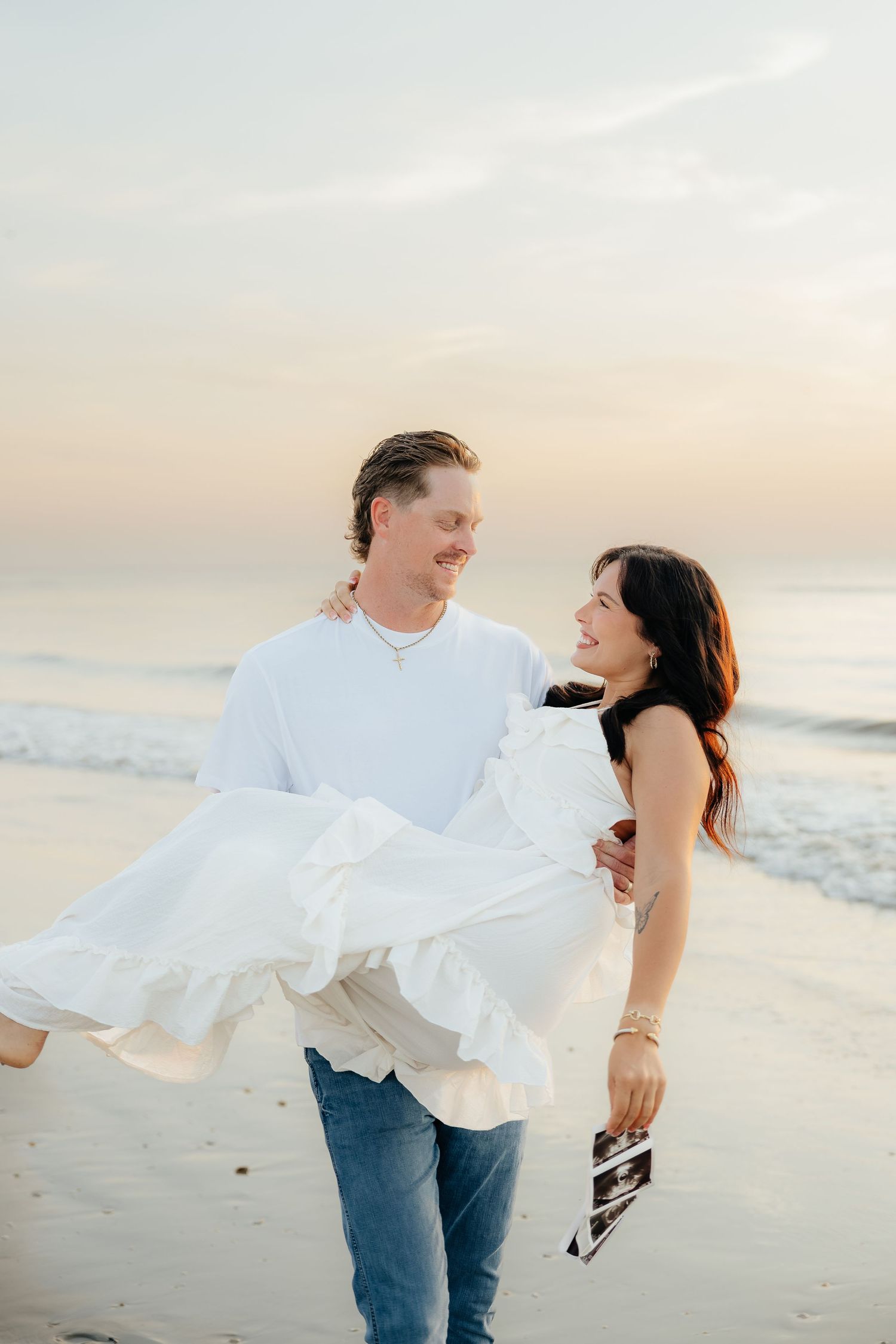 A couple shares a joyful moment on the beach, man carrying woman in white dress, holding an ultrasound photo.
