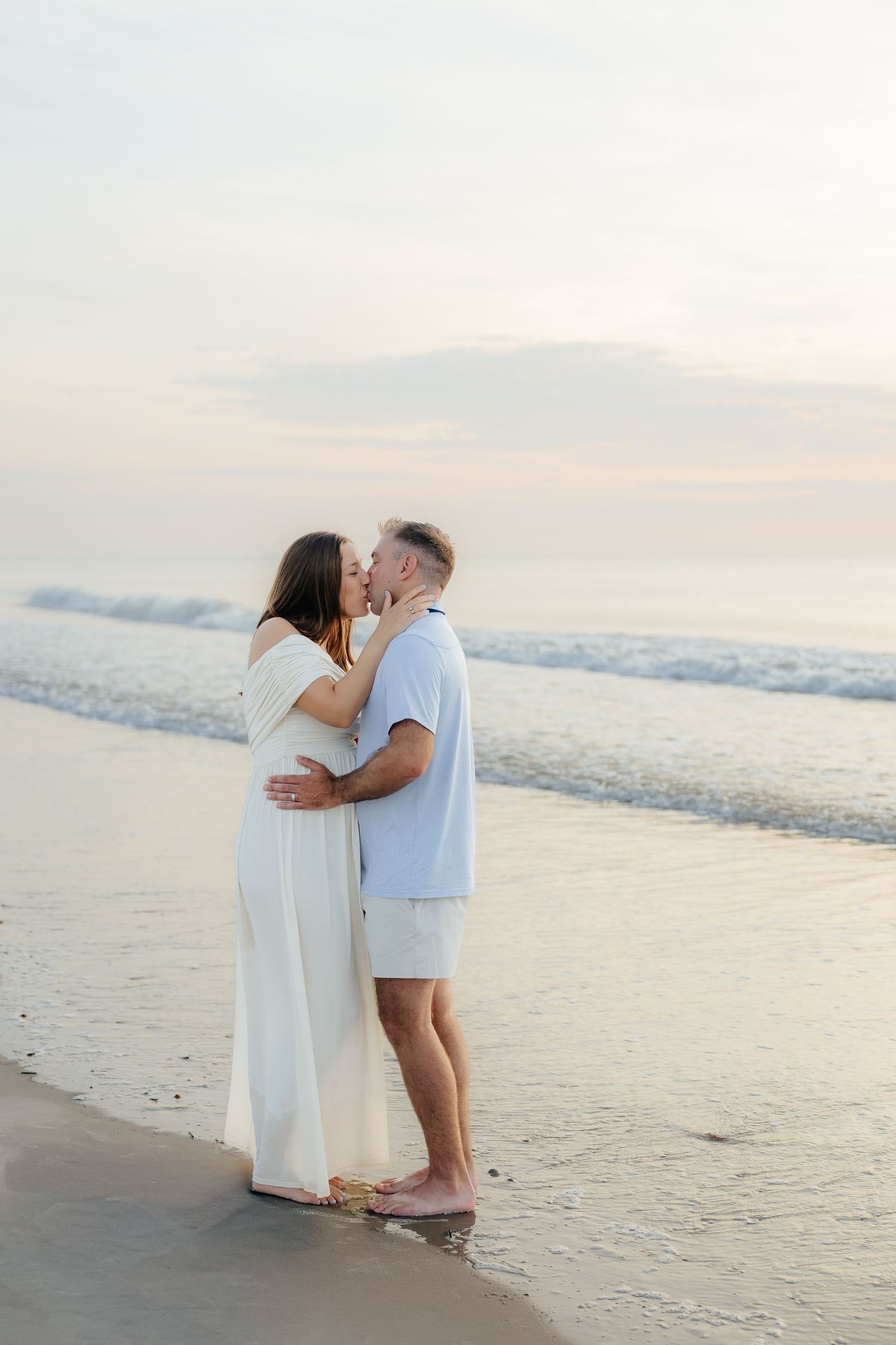 A couple shares a romantic kiss on the beach at sunset, dressed in white and light blue outfits with waves behind them.