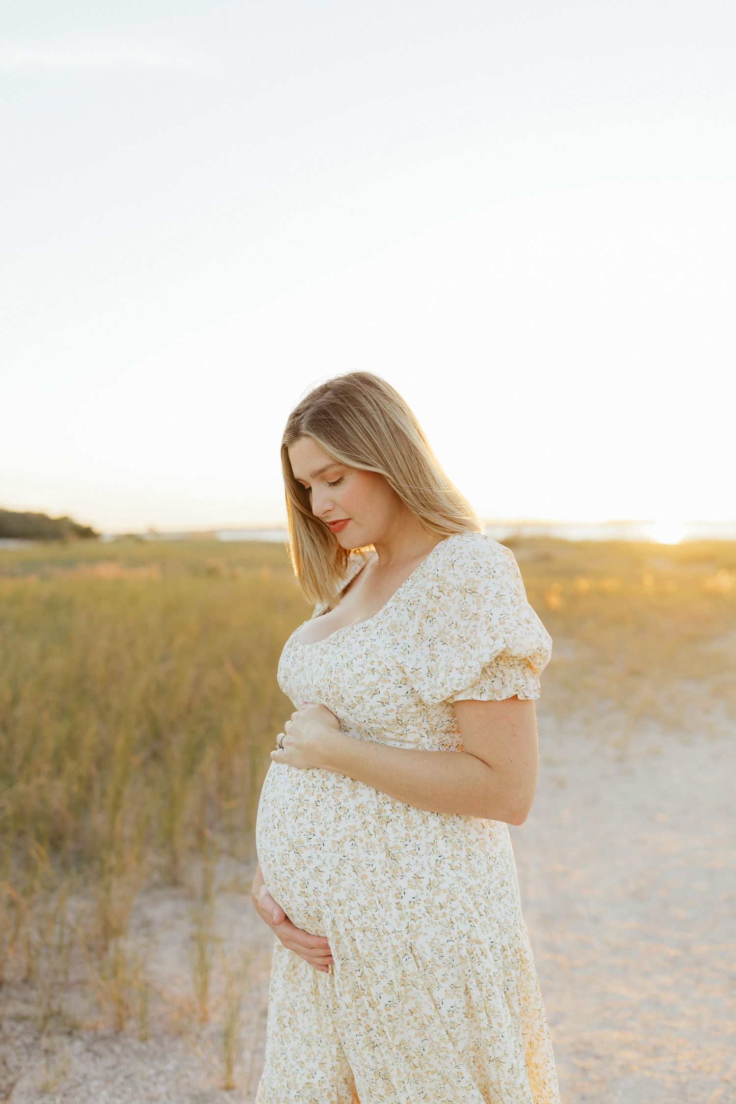 Expectant mother in a floral dress cradles her baby bump during golden hour at the Fort Clinch State Park beach.