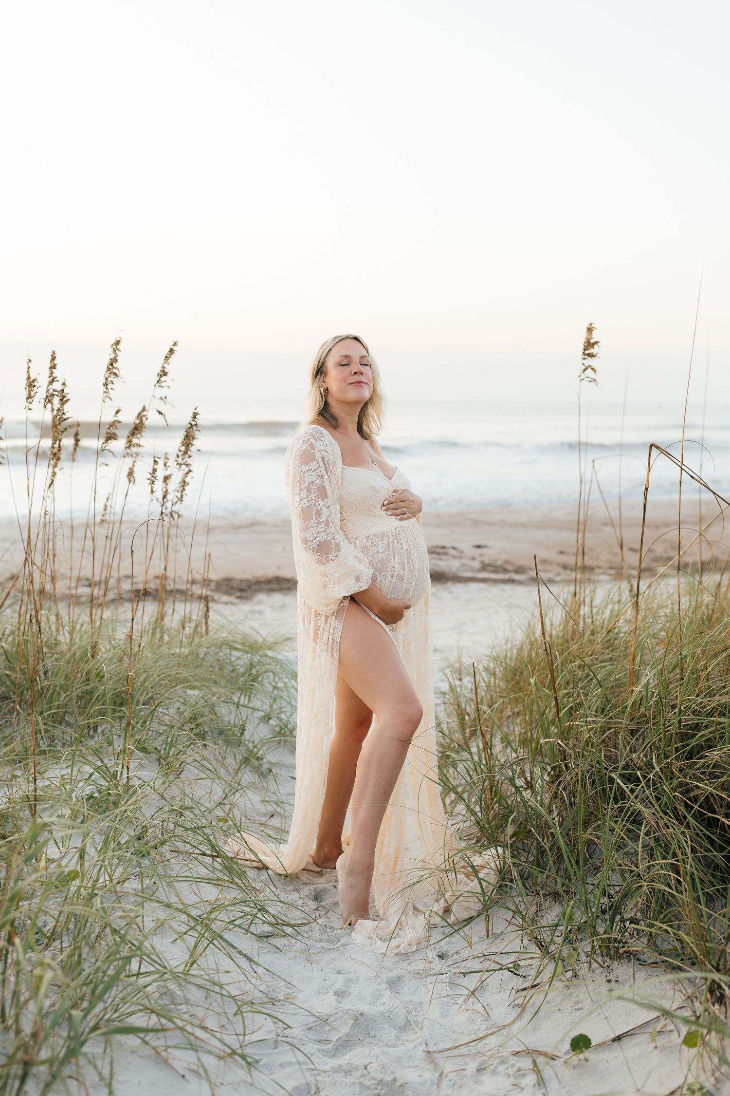 Expectant mother in lace dress poses among beach grass dunes at sunset.