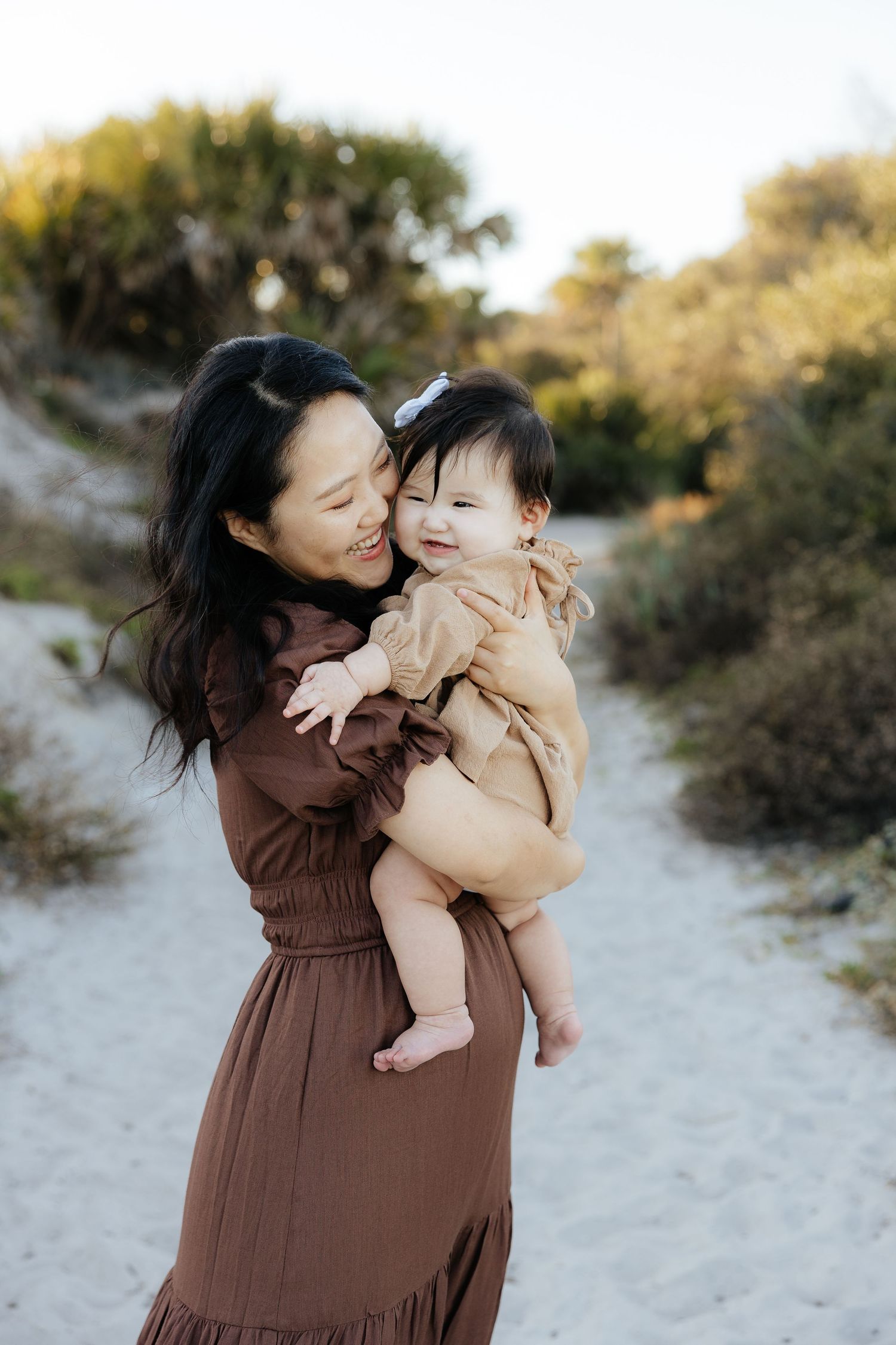 A parent in a brown dress shares joyful moments with a baby on a sandy path surrounded by greenery.