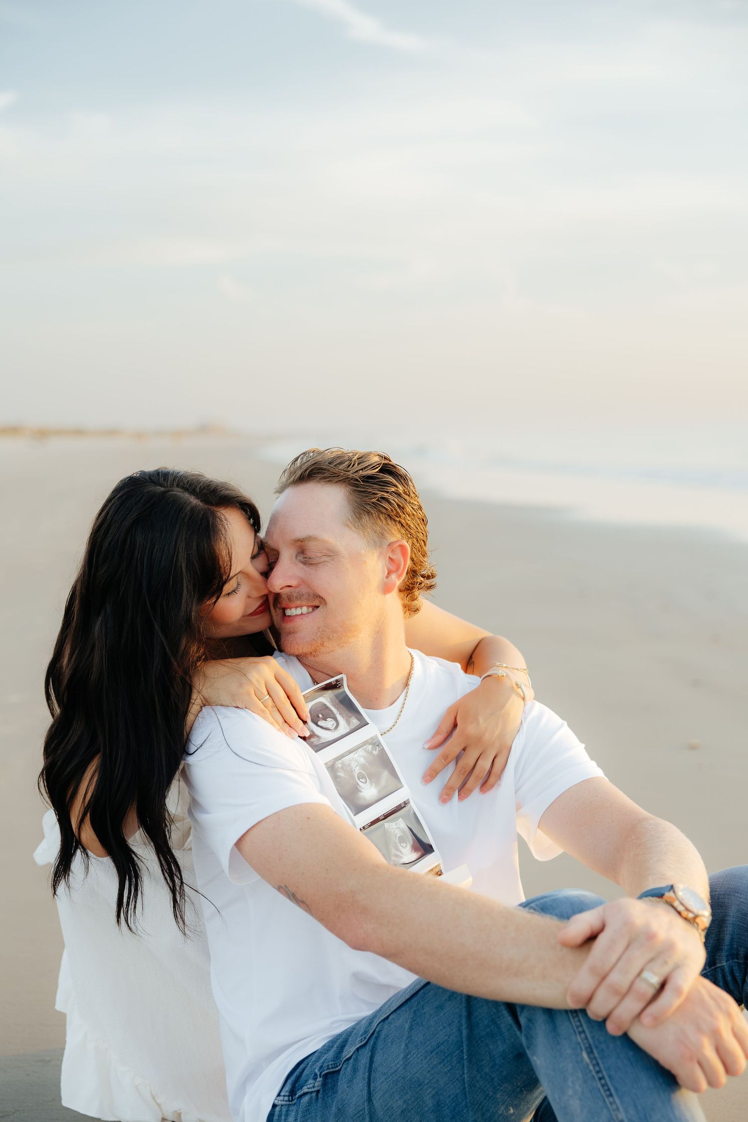 Happy couple sitting on beach, woman kissing man's cheek as he holds ultrasound photos, pregnancy announcement photo.