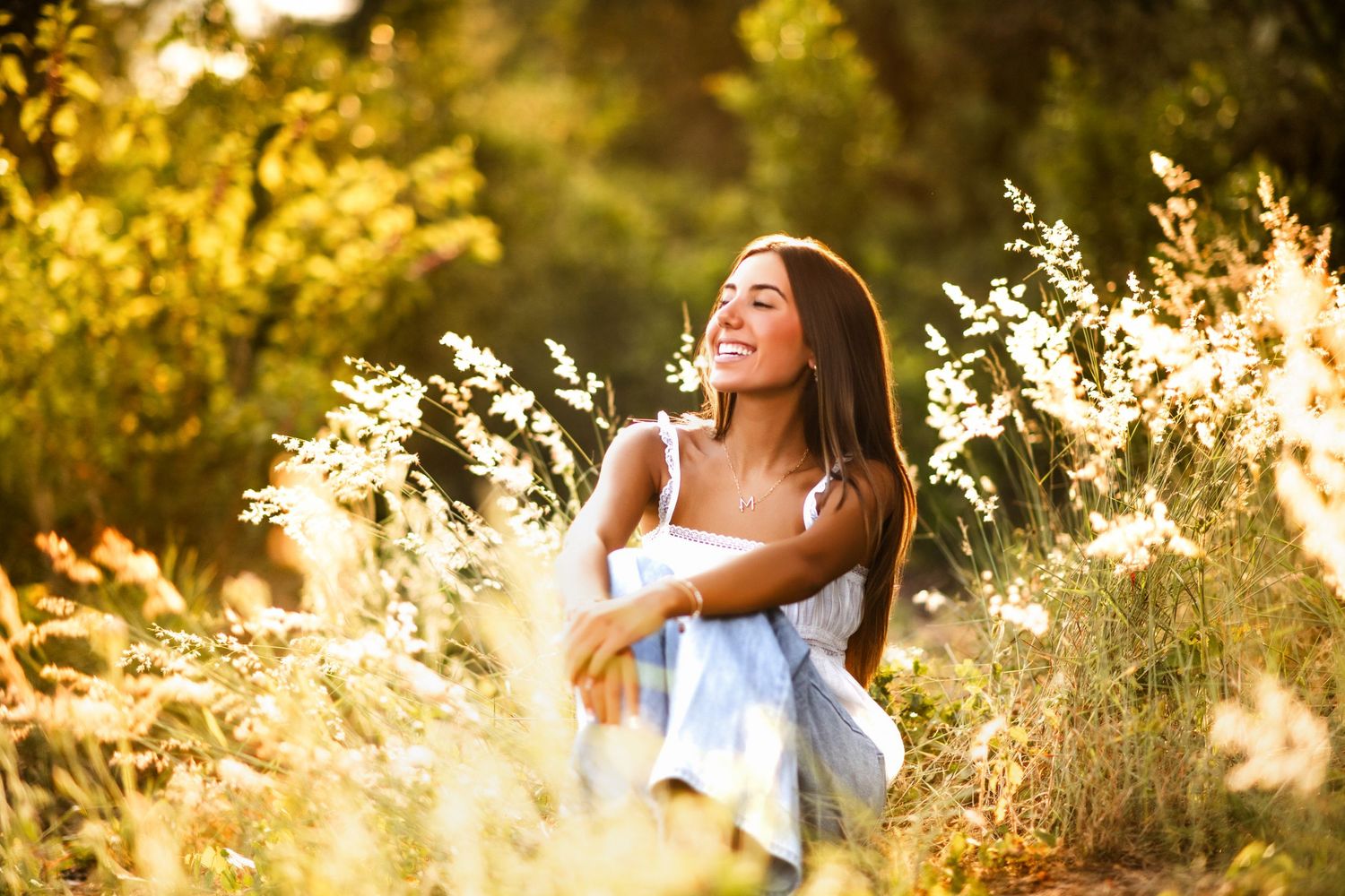 A smiling young woman sits outdoors among golden wildflowers and sunlit trees on a warm sunny day.