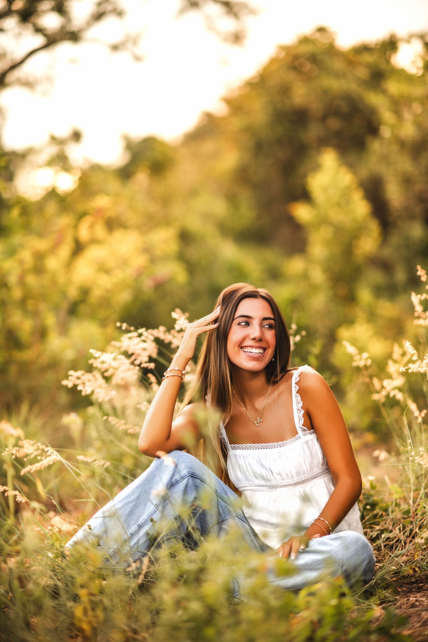 Young woman in white top and jeans sitting in a sunlit meadow, smiling warmly among wildflowers.