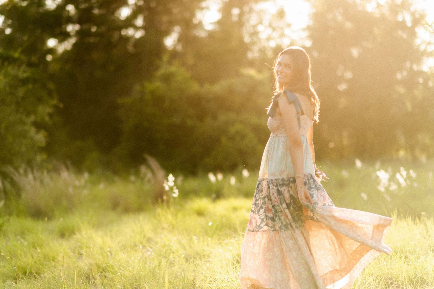 Young woman in floral dress twirling in sunlit meadow with golden hour light and lush green trees.