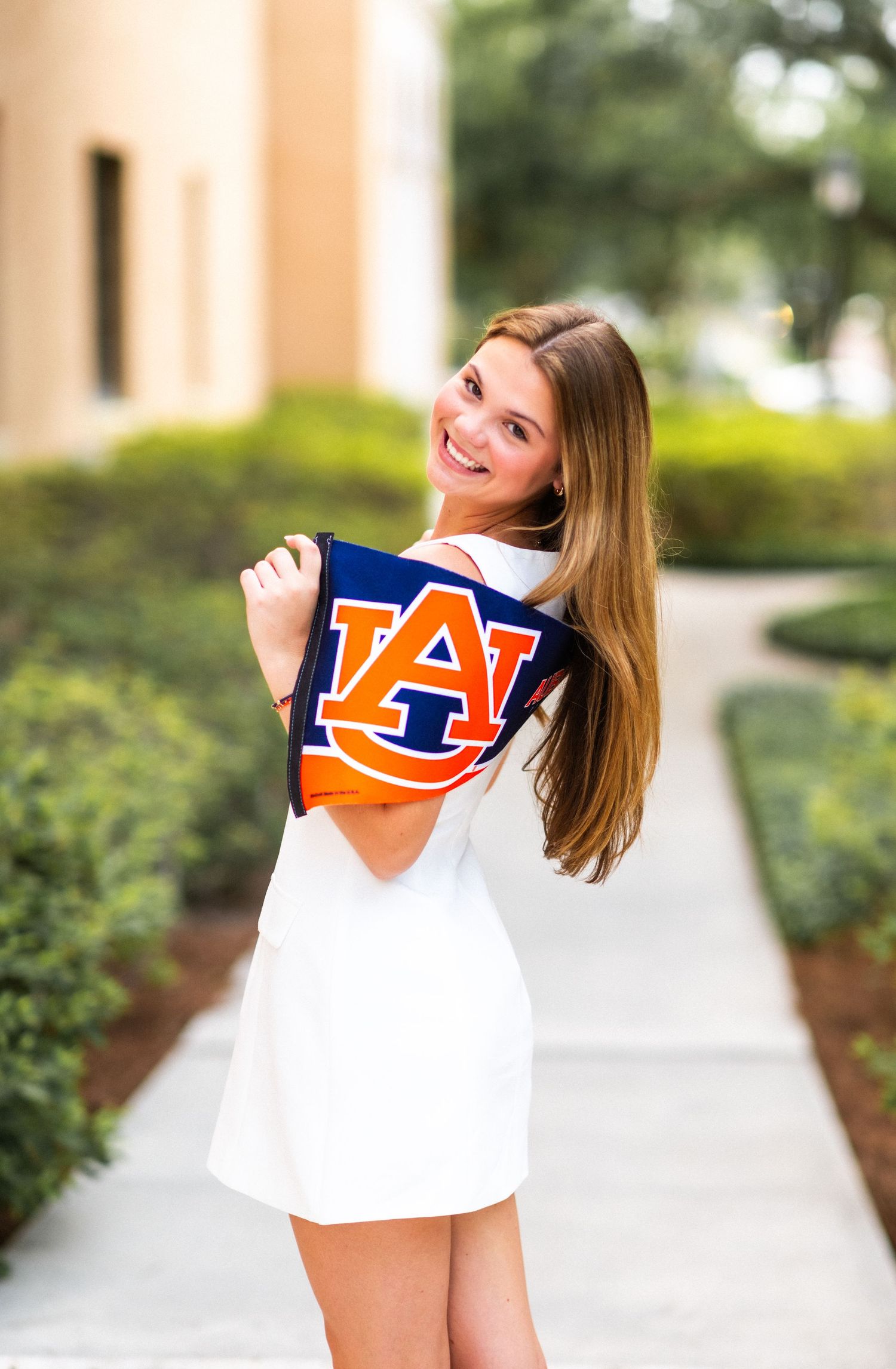 Smiling young woman in white dress holding Auburn University AU flag pennant on a sunny campus walkway.