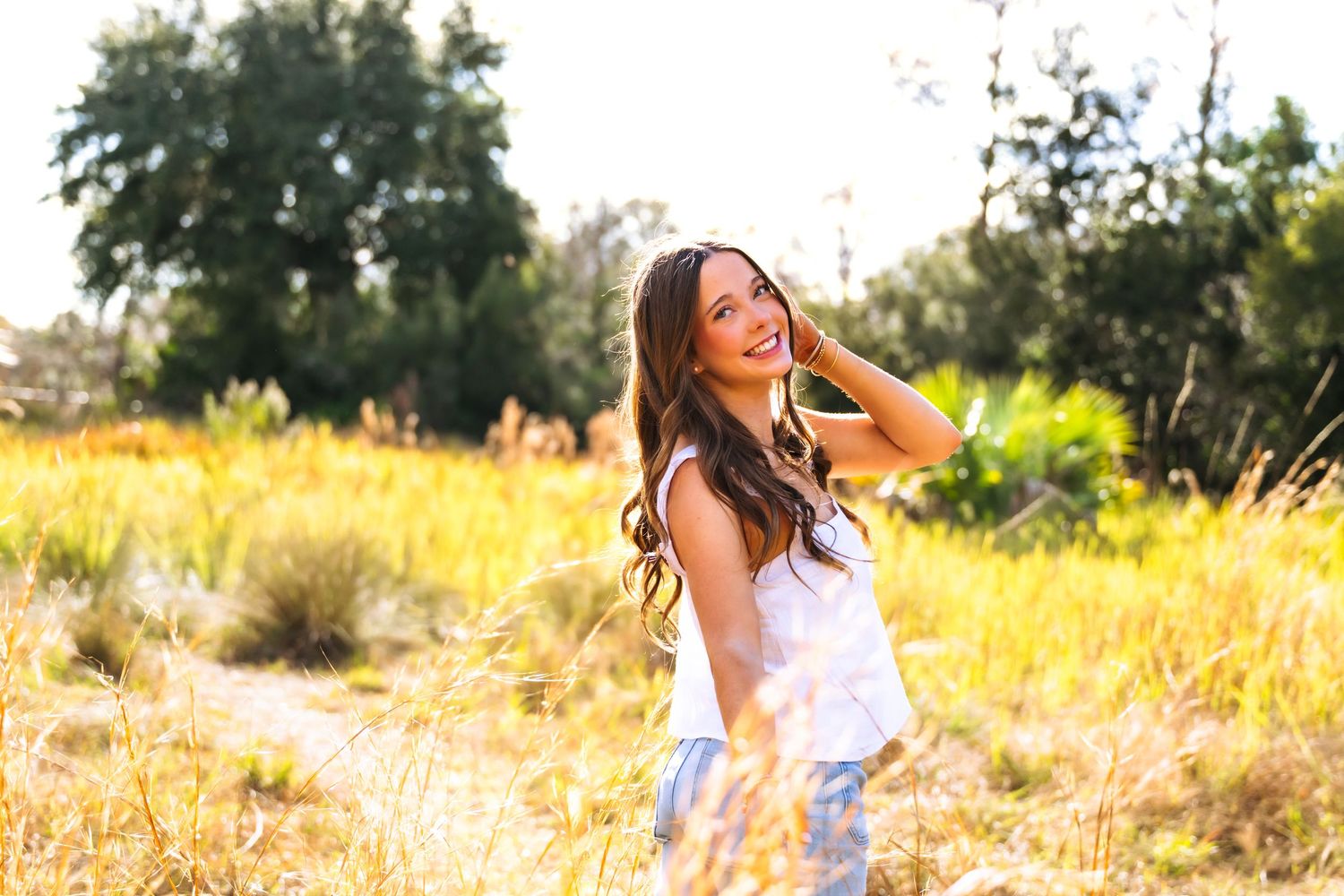 Young woman with long dark hair smiling in a sunlit golden field, wearing a white top on a warm sunny day.