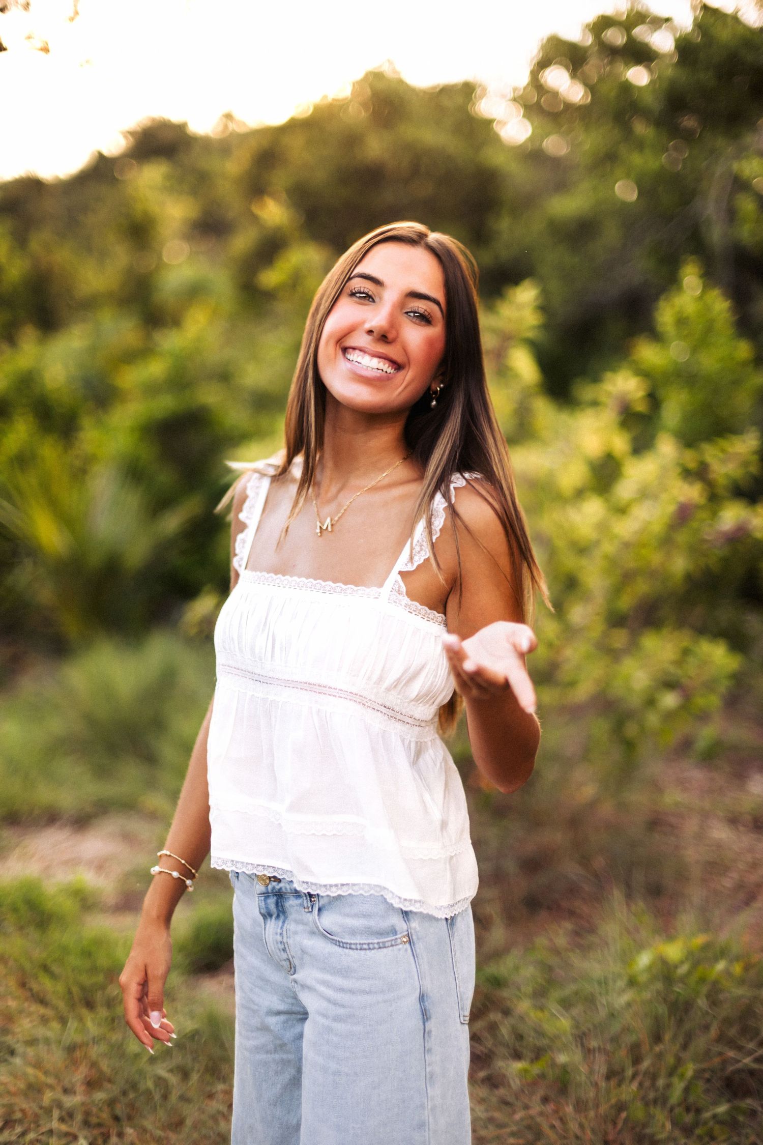 Smiling young woman in white top and jeans reaching out hand in sunlit green nature setting.