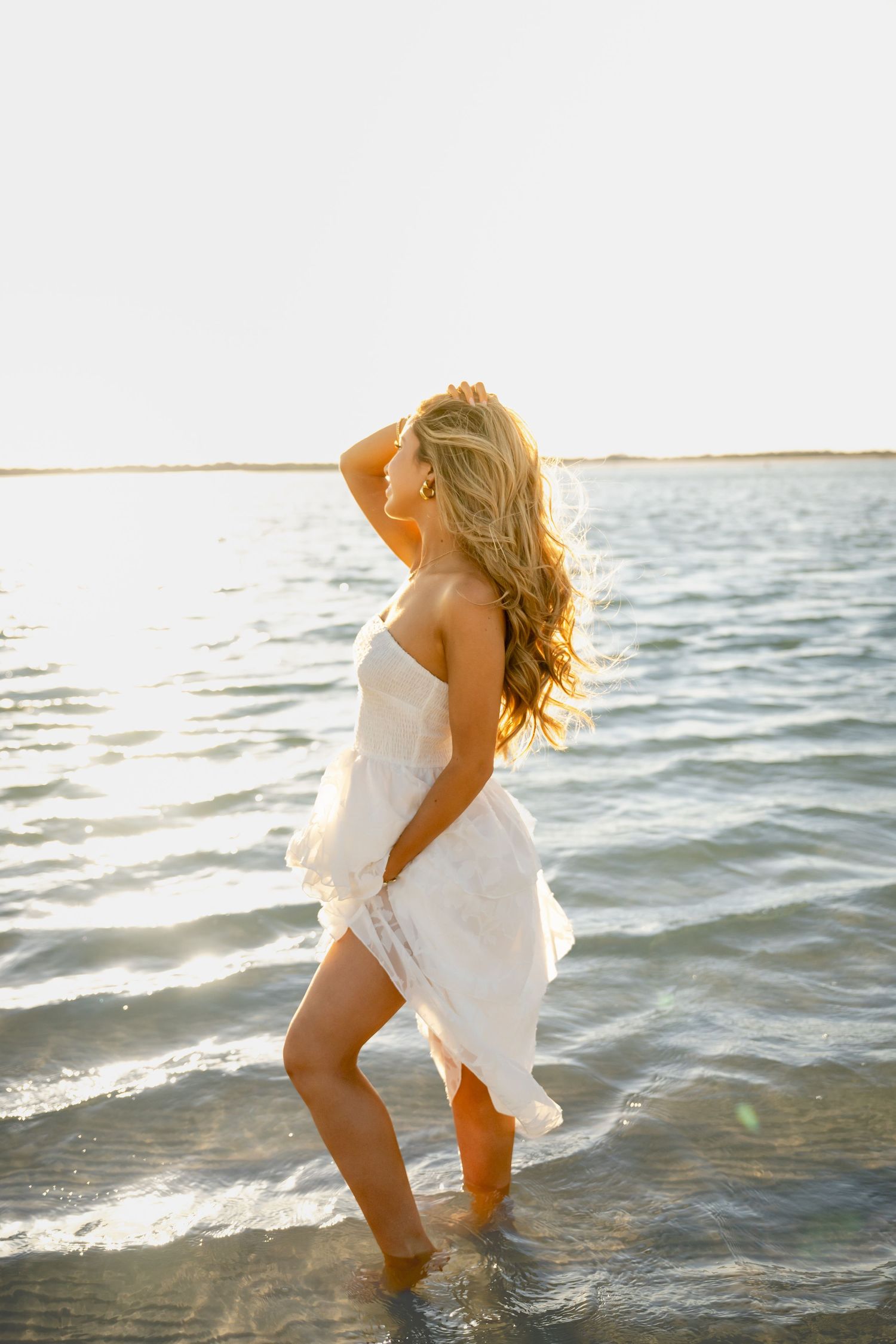 Woman in white dress standing in shallow ocean water at golden hour with long blonde hair blowing in the breeze.