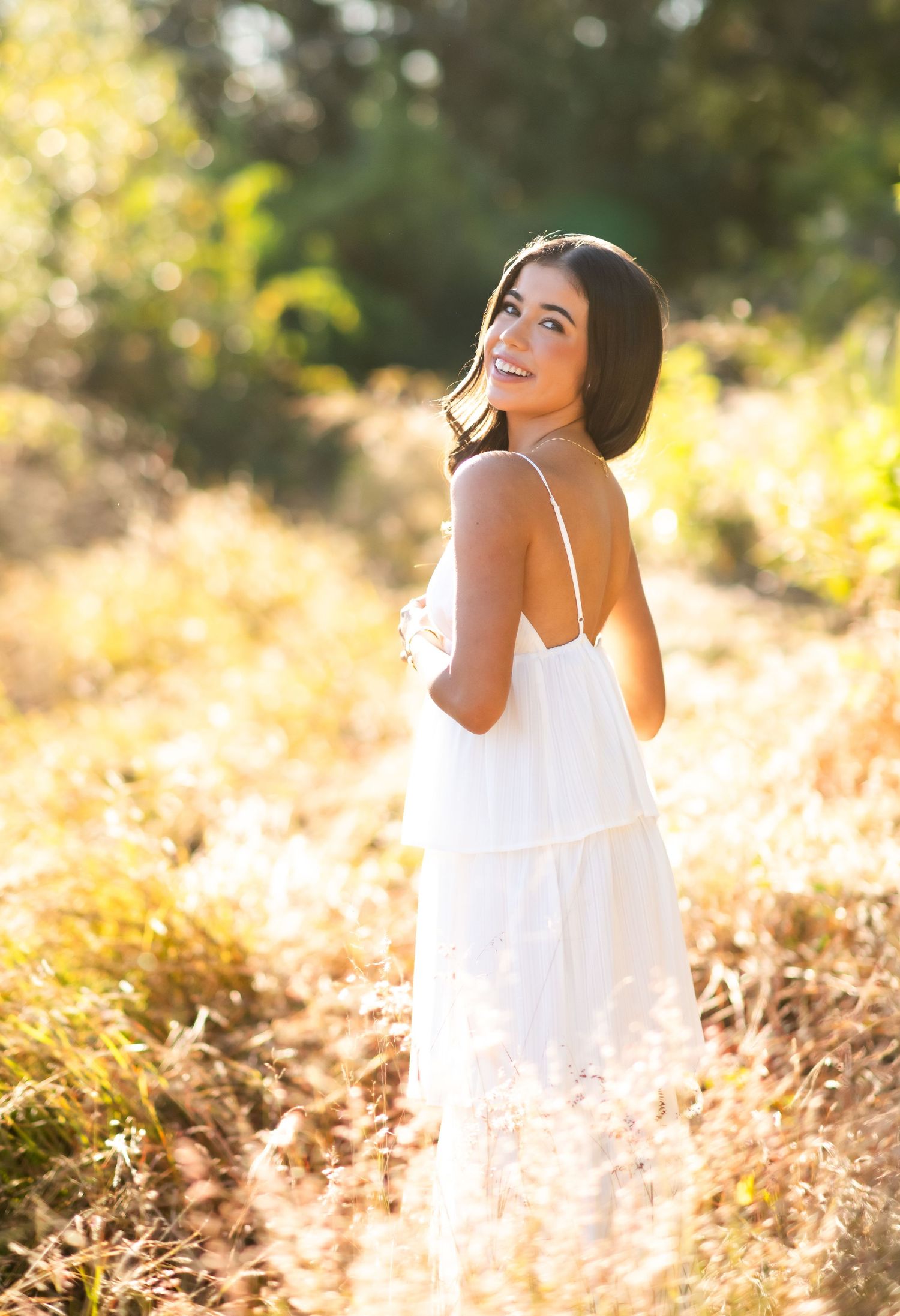 Smiling young woman in white sundress standing in golden sunlit field, glancing over shoulder.